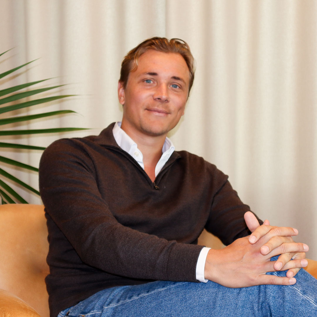 Man med ljusbrunt hår, klädd i svart tröja och blå jeans, sitter korslagd i en orange stol med händerna samlade.