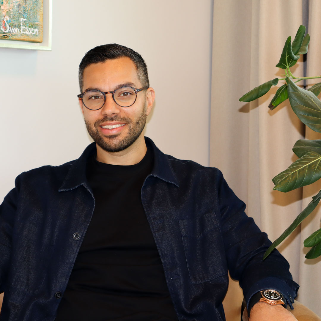 Man med glasögon och mörk skjorta sitter leende framför en beige bakgrund med gröna växtblad till höger.