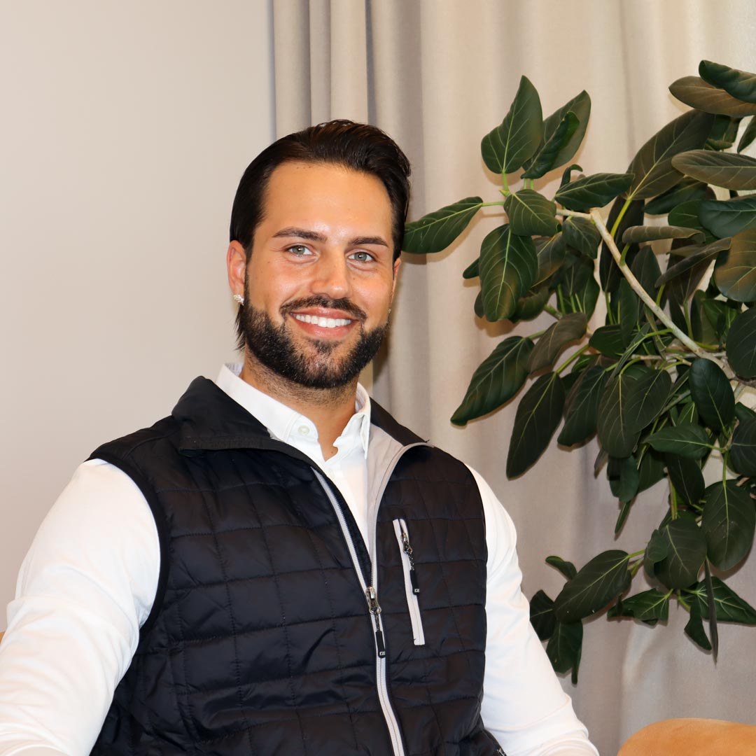 Leende man med skägg och svart väst sitter framför en växt med gröna blad.