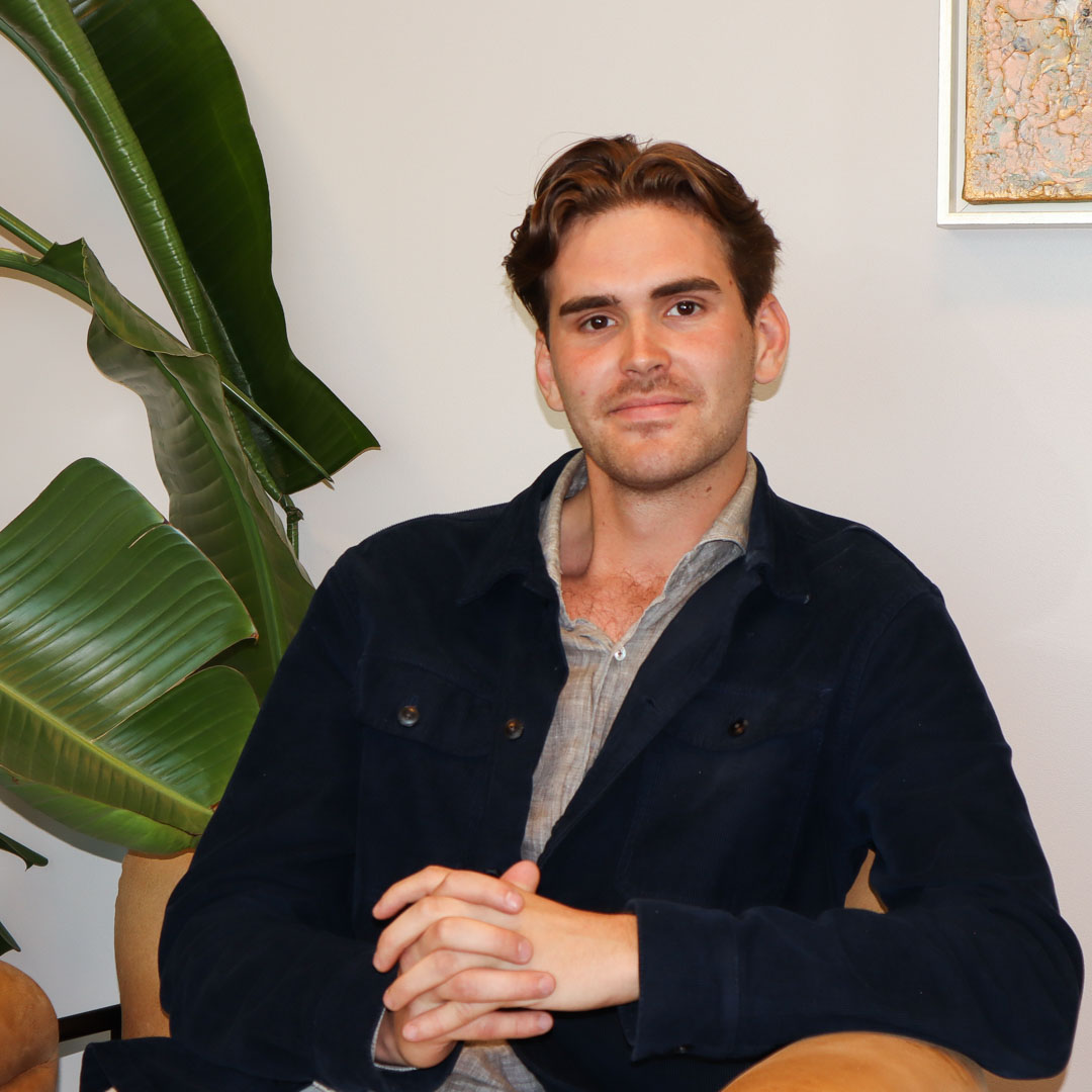 Ung man med mörkt hår i mörk jacka sitter avslappnat med händerna ihop i en fåtölj, med stora gröna blad i bakgrunden.