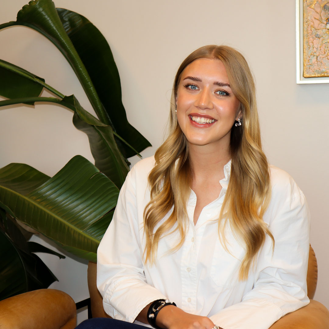 Leende kvinna med långt blont hår och vit skjorta, sittande i en fåtölj med stora gröna växtblad i bakgrunden.