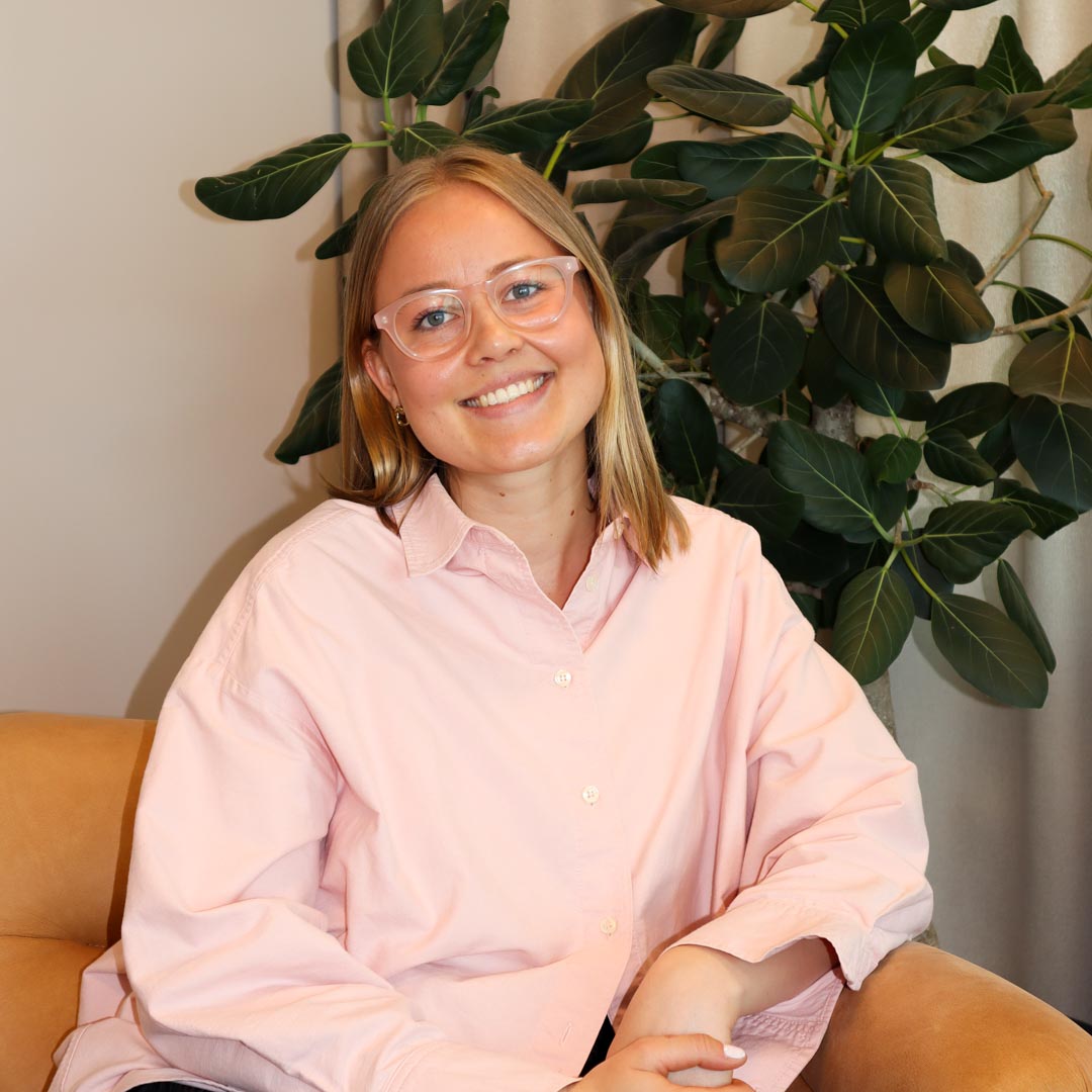 Kvinna med glasögon och ljusrosa skjorta sitter leende i en fåtölj med en stor grön växt i bakgrunden.