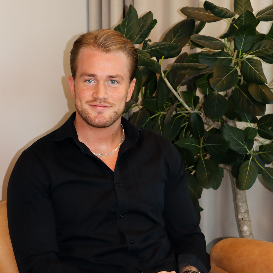Ung man med ljusbrunt hår och ljusblå ögon klädd i svart skjorta sitter i en stol framför en grön växt.