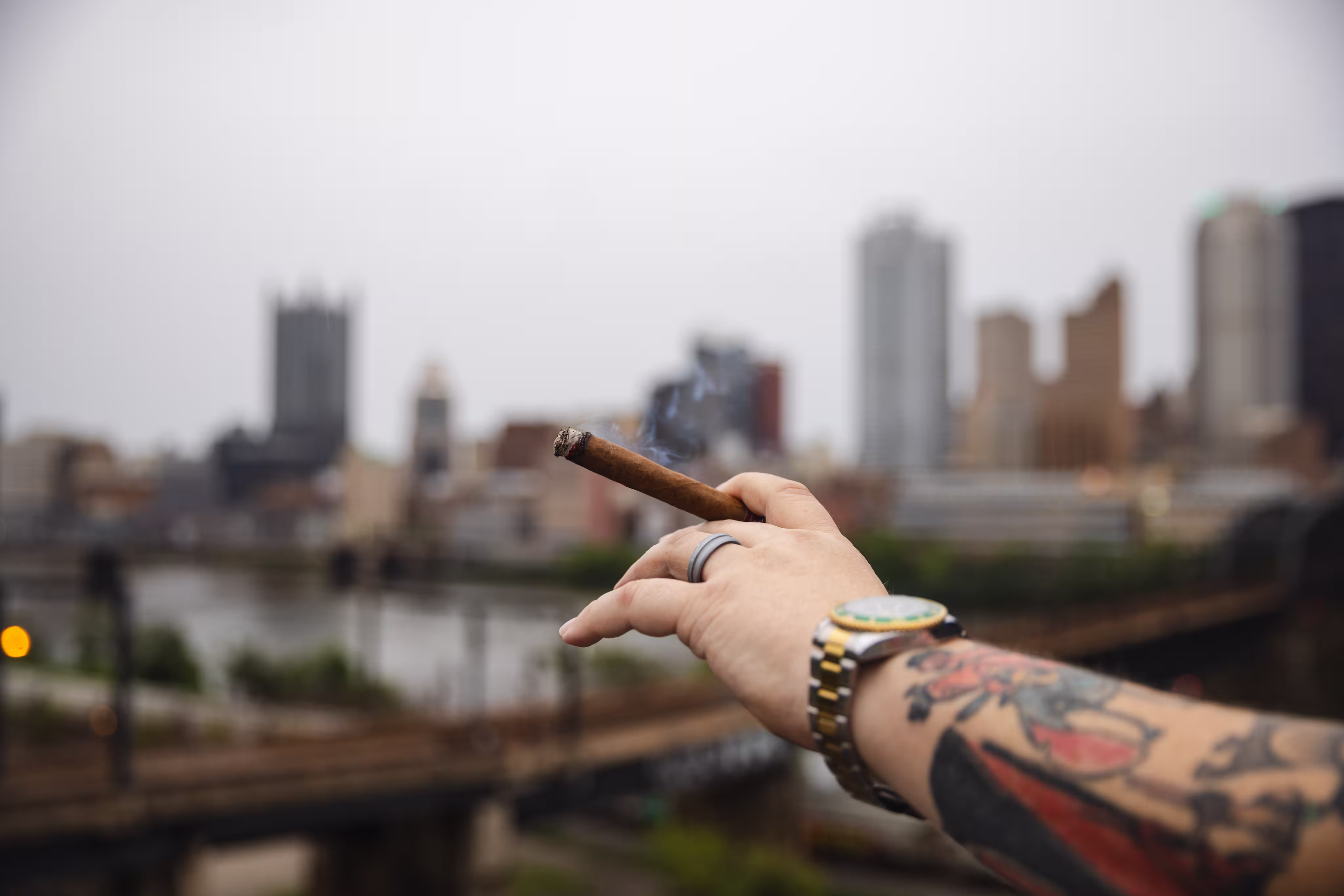 Cigar held over railing at The Distillery Complex with background of extraordinary Downtown Pittsburgh skyline view. Photo by Alex Mowery