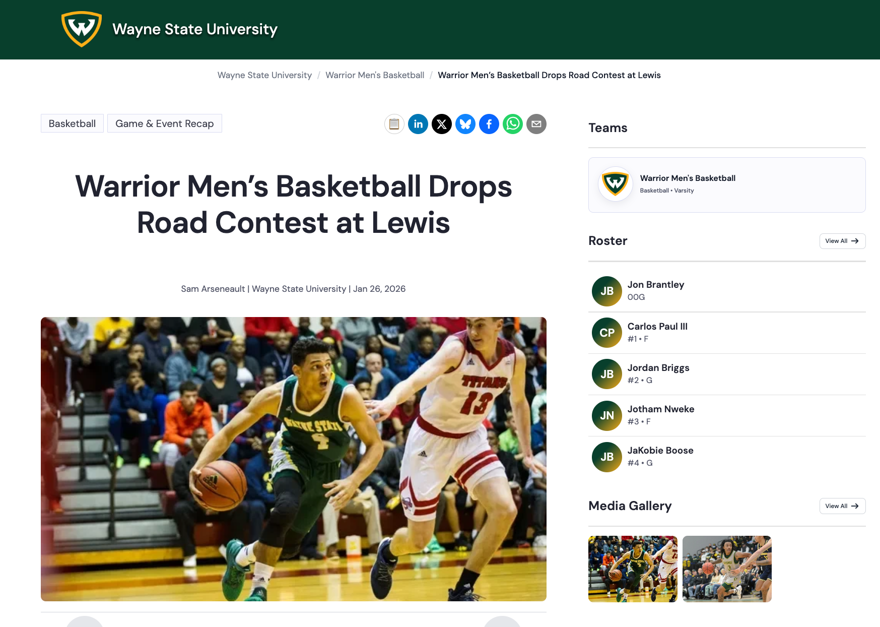 Wayne State basketball player dribbling the ball while being defended by a Lewis player in a gymnasium with spectators in the background.