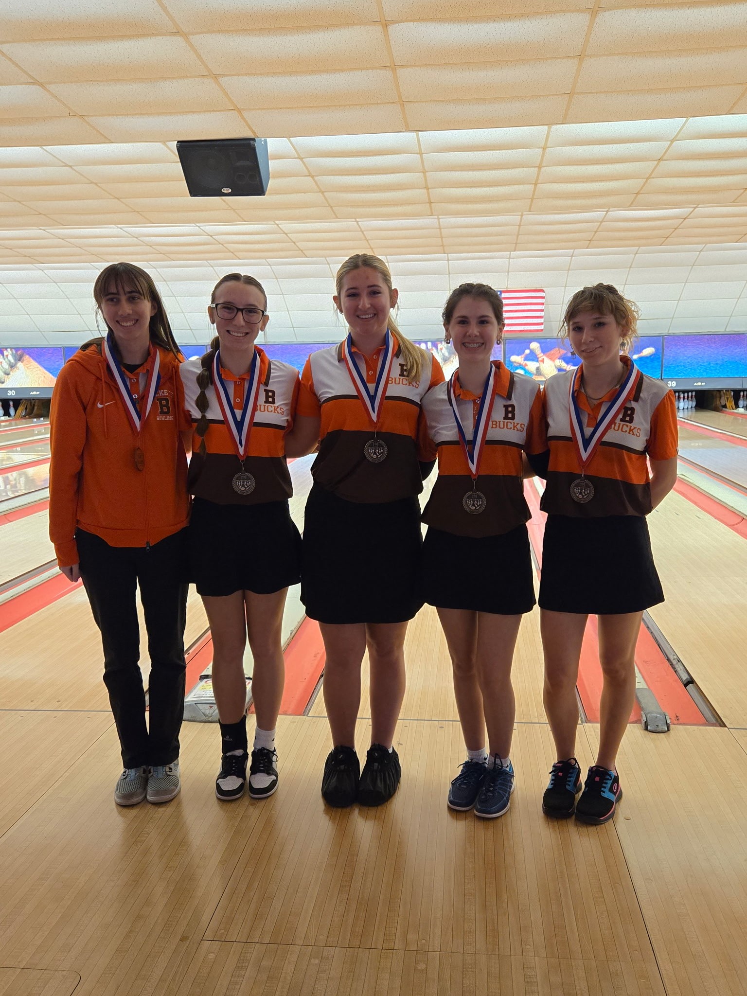 Five female high school bowlers standing in a bowling alley wearing medals, four in team uniforms and one in an orange jacket.