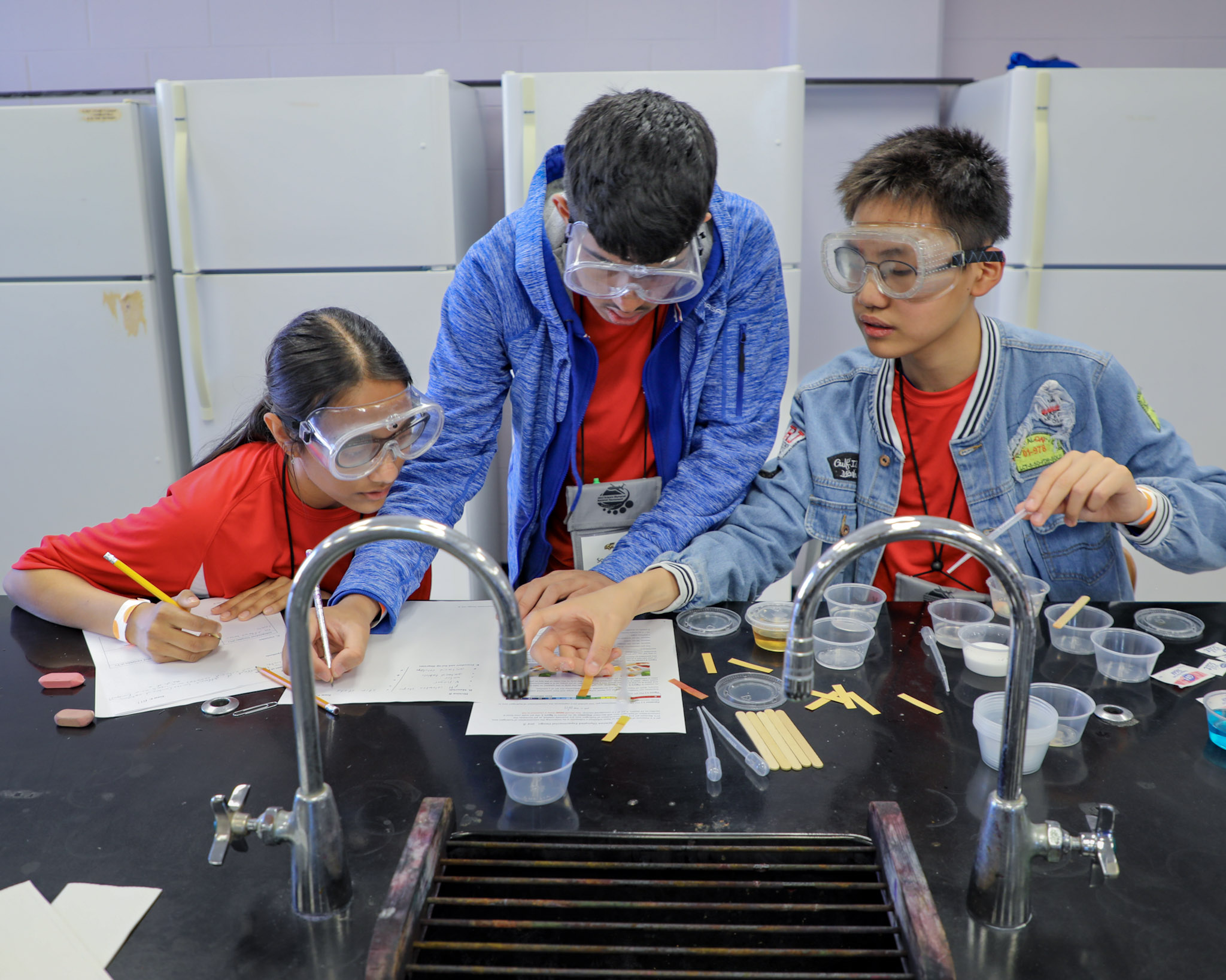 Three students in safety goggles conducting a science experiment with pH test strips and liquids in clear cups at a lab table.