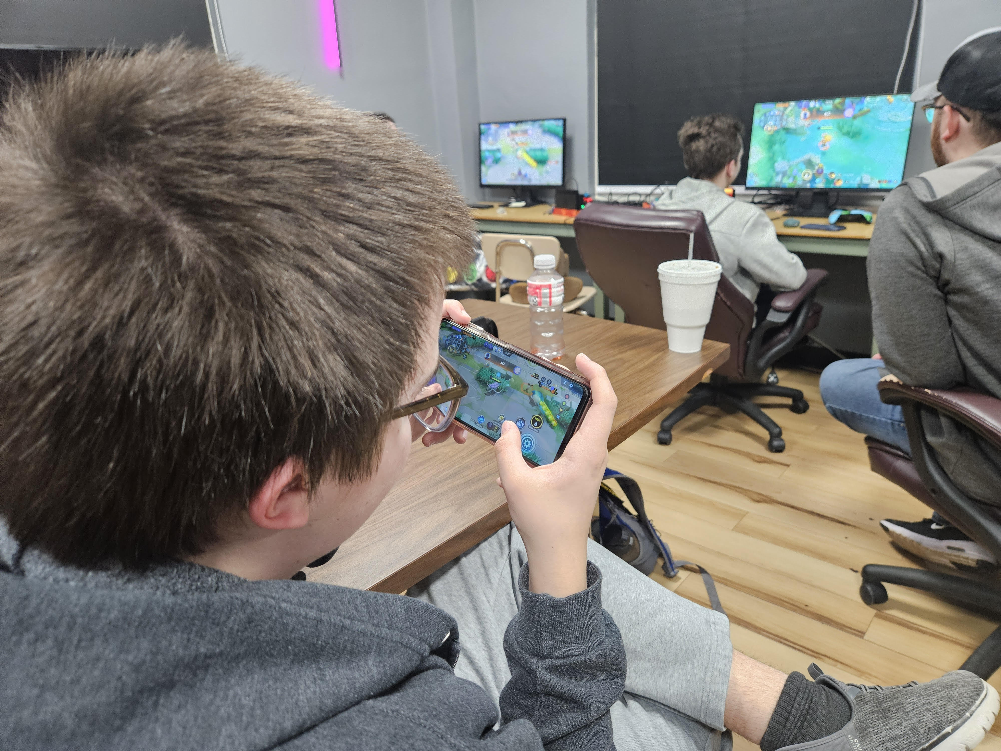 Teen boy wearing glasses and gray sweatshirt playing a game on a smartphone, with two people playing video games on monitors in the background in a casual room.