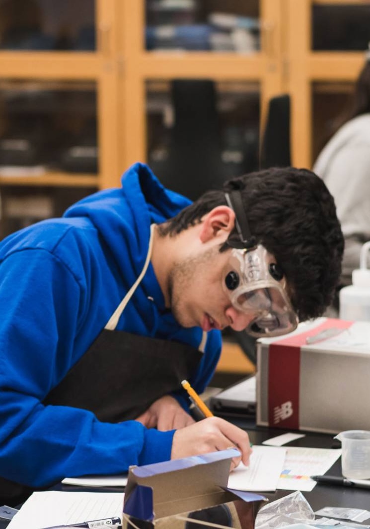 Student wearing safety goggles and a blue hoodie writing with a pencil on paper in a classroom.