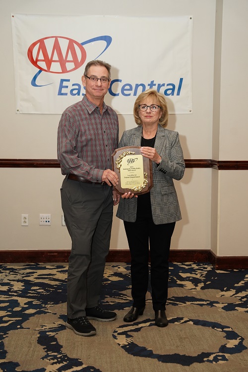 Two people standing indoors holding a plaque in front of a banner that reads AAA East Central.