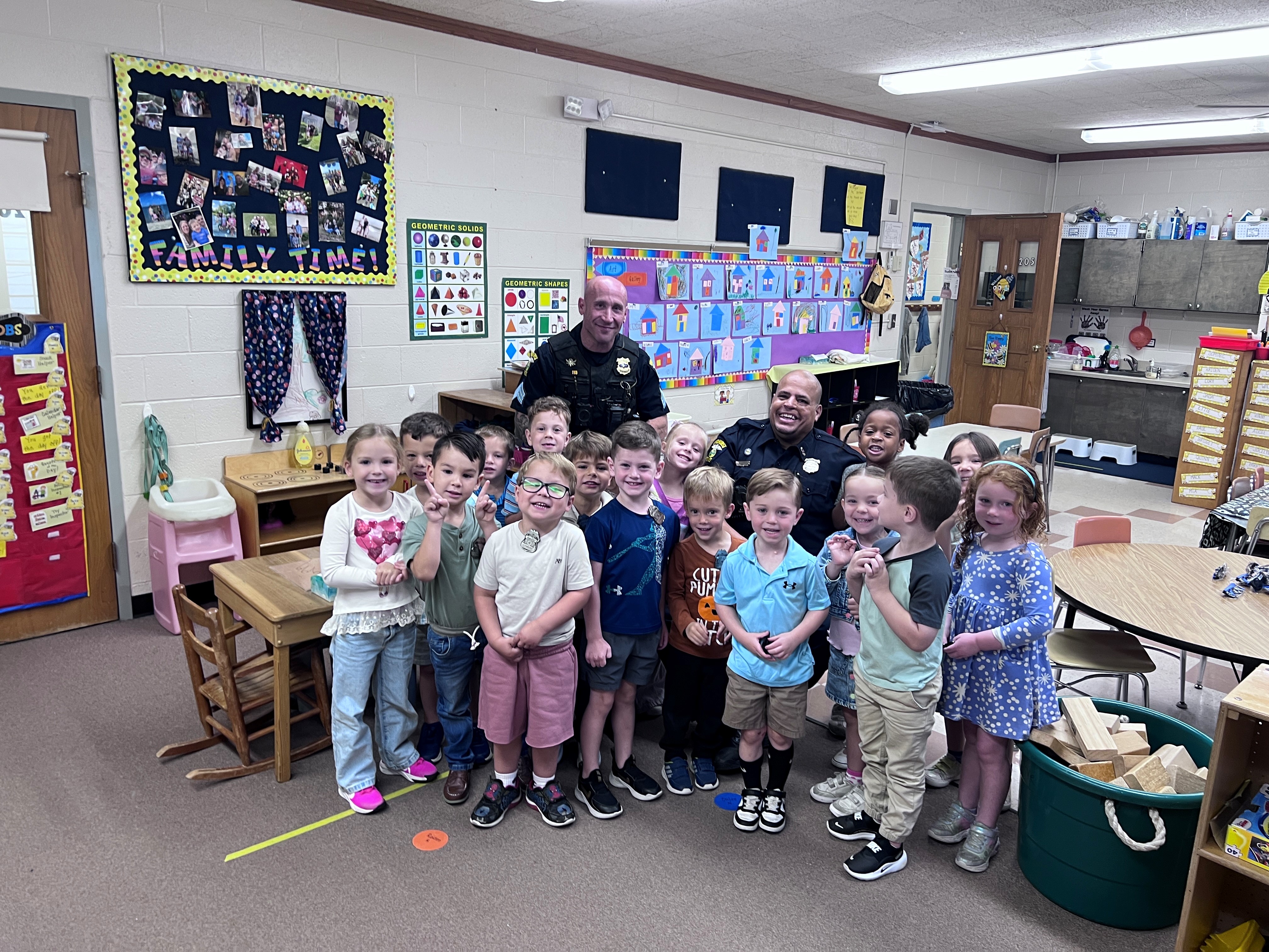 Group of smiling young children posing with two police officers in a colorful classroom.