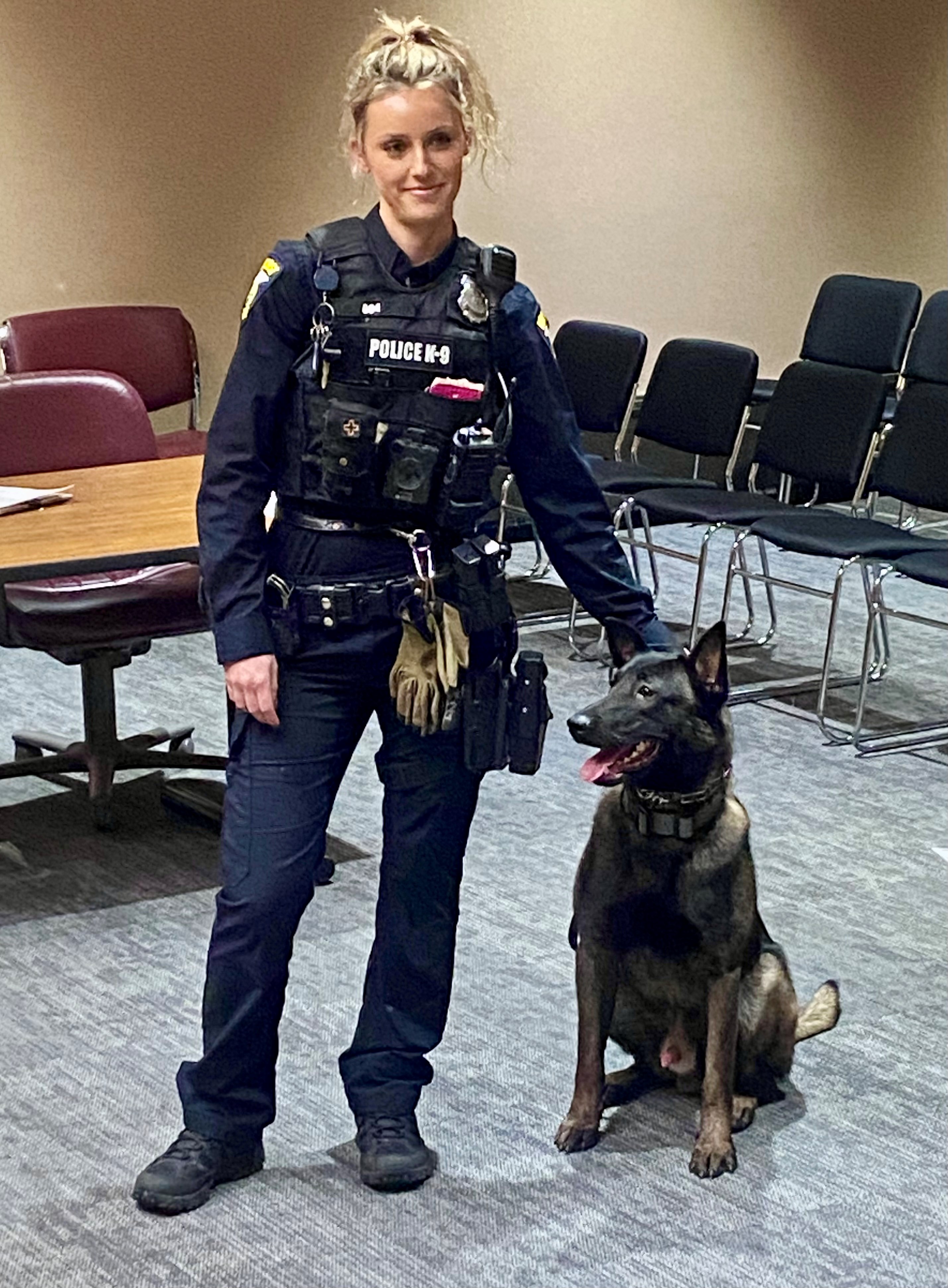 Female police officer in K-9 uniform standing indoors with a sitting police dog.