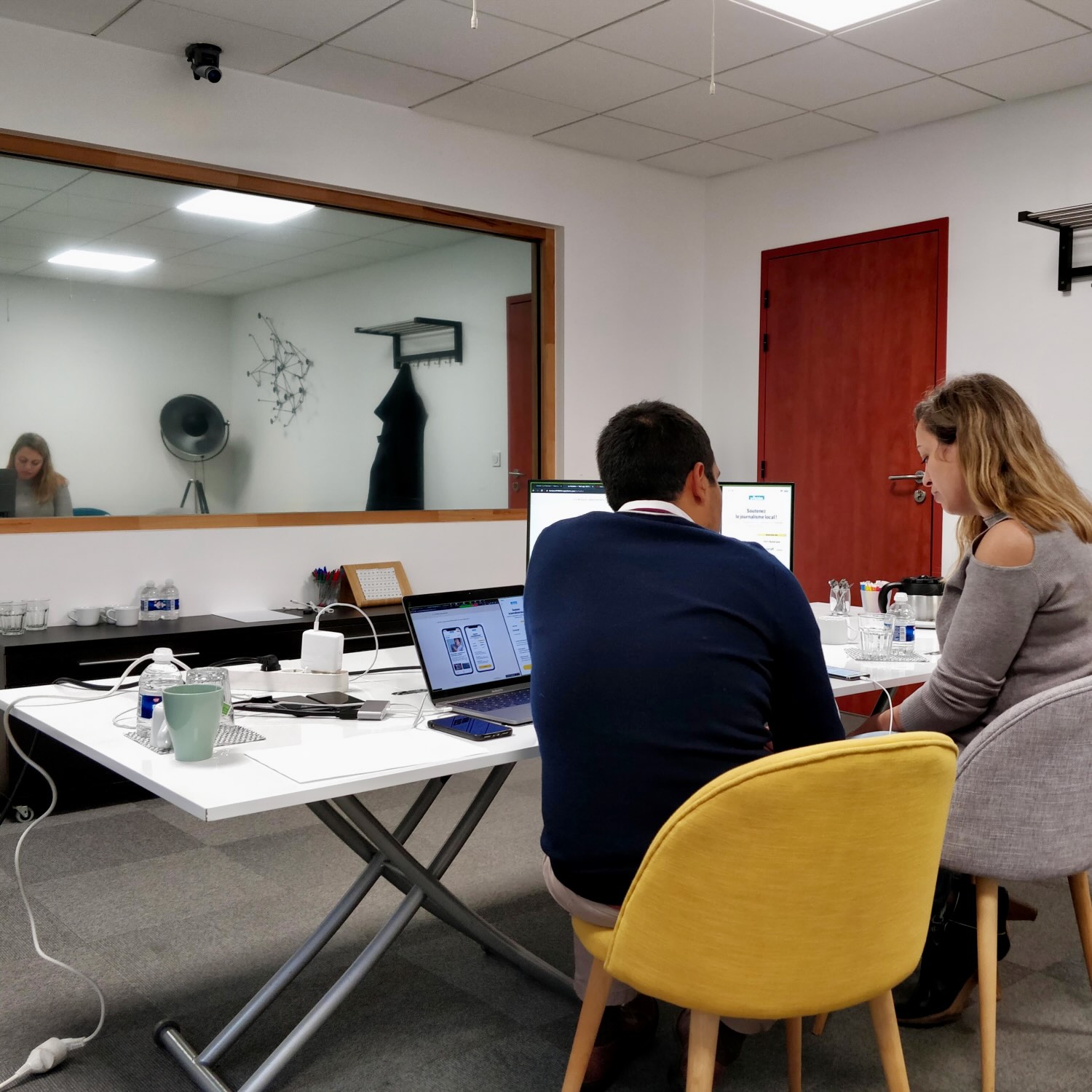 Two people seated at a white table with laptops and a monitor in a modern office room with grey carpet and a large window reflecting another person.