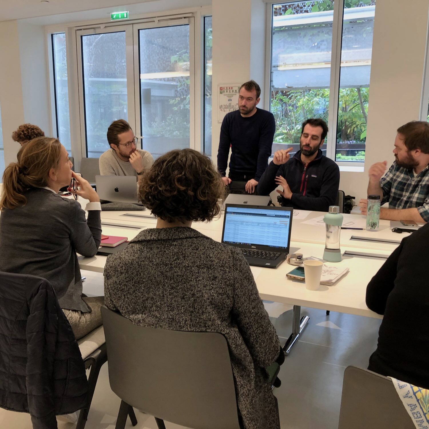 Group of six people engaged in a meeting around a table with laptops, papers, and water bottles in a bright room with large windows.
