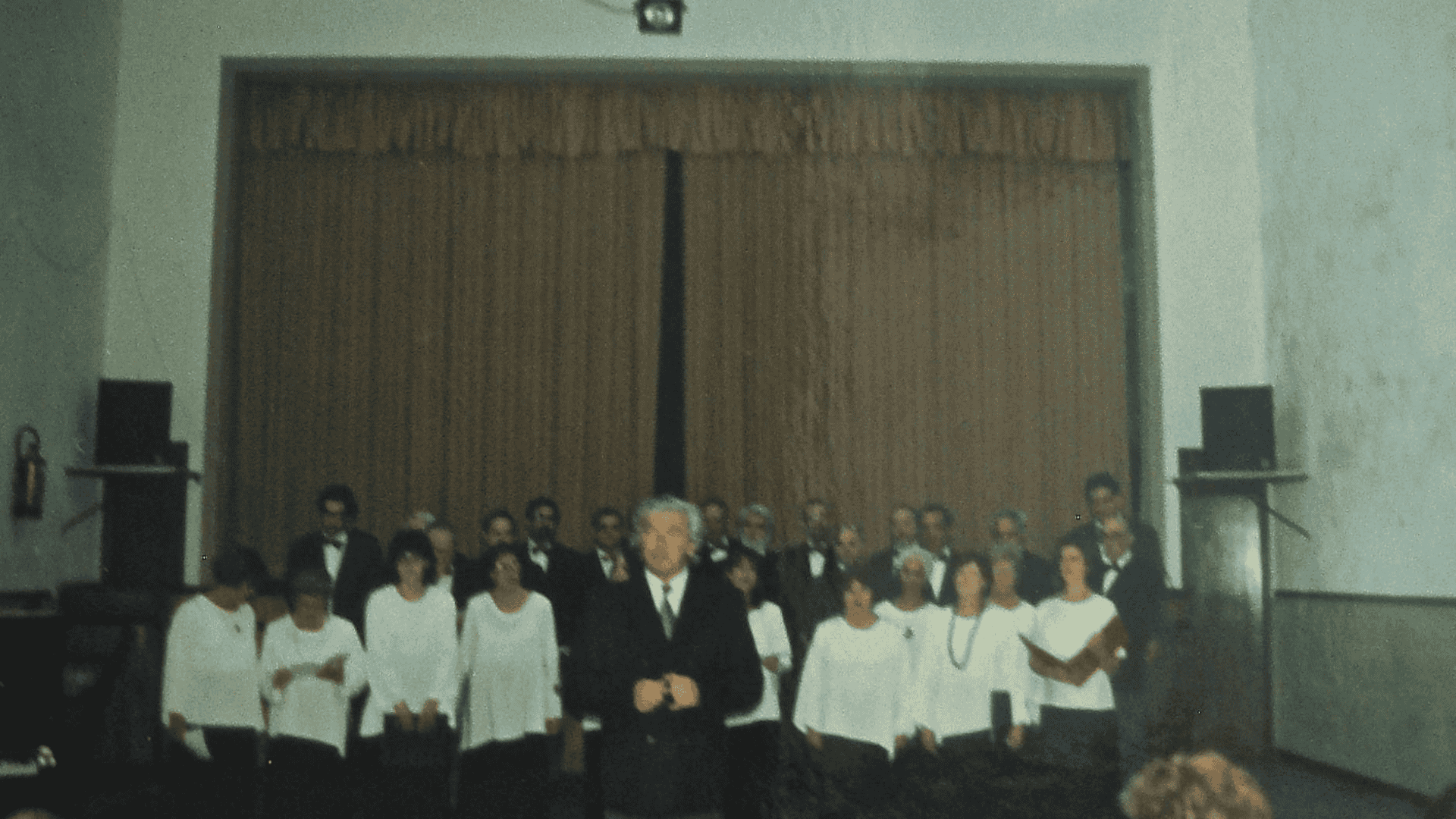 Foto antiga do Salão de Espetáculos da SFUA, com o coro e o maestro em cena prontos para cantar