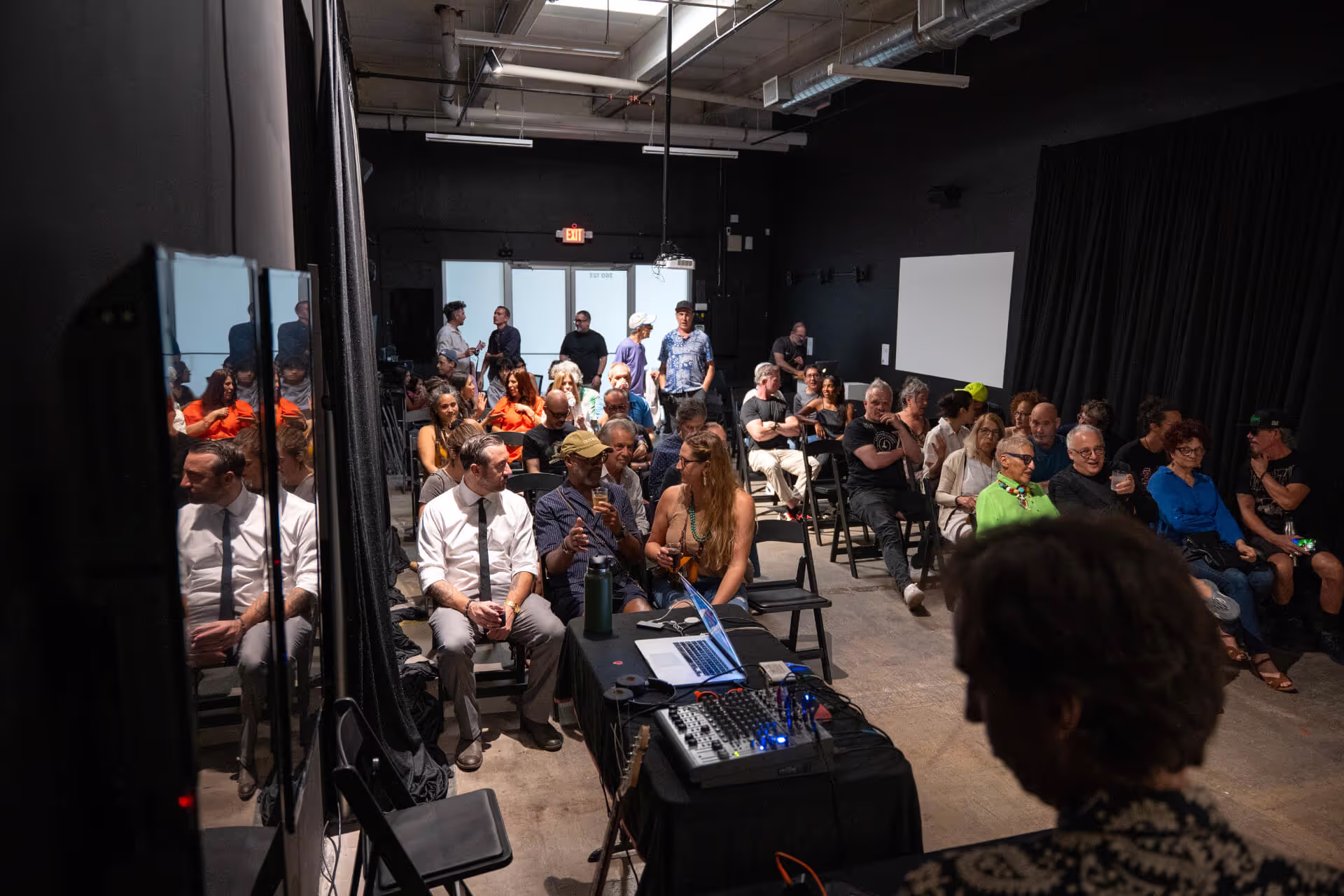 MUD Foundation The Sound of Art Experimental Music and Sound Art Program, featuring an audience seated in a dimly lit room while a person adjusts sound equipment and a laptop at the front of the space.