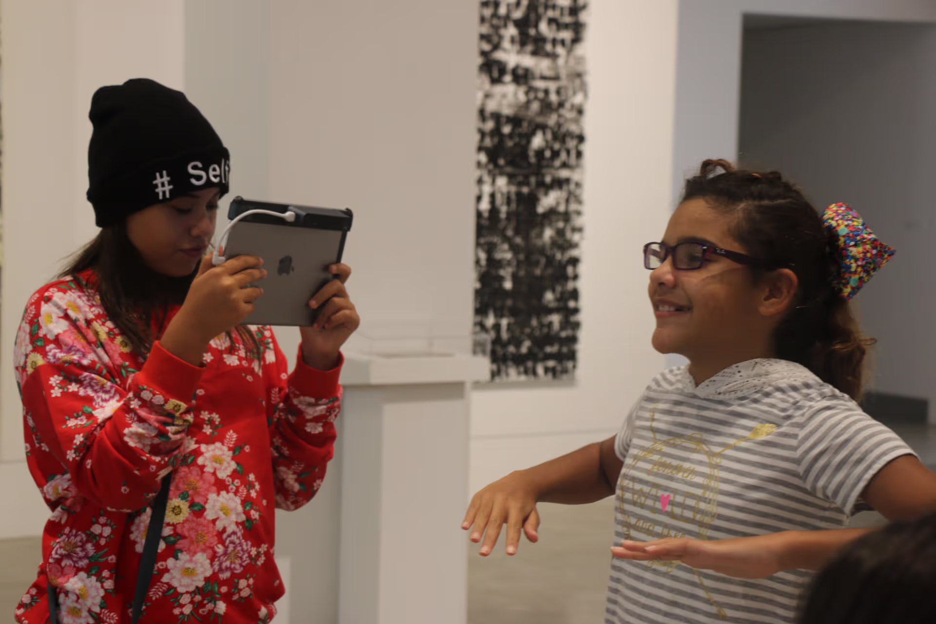 MUD Foundation XRCamp at the De La Cruz Collection in Miami, Florida, 2018, featuring a girl holding a tablet and photographing another girl wearing glasses and a striped shirt, smiling with her arms outstretched in a minimalist room.