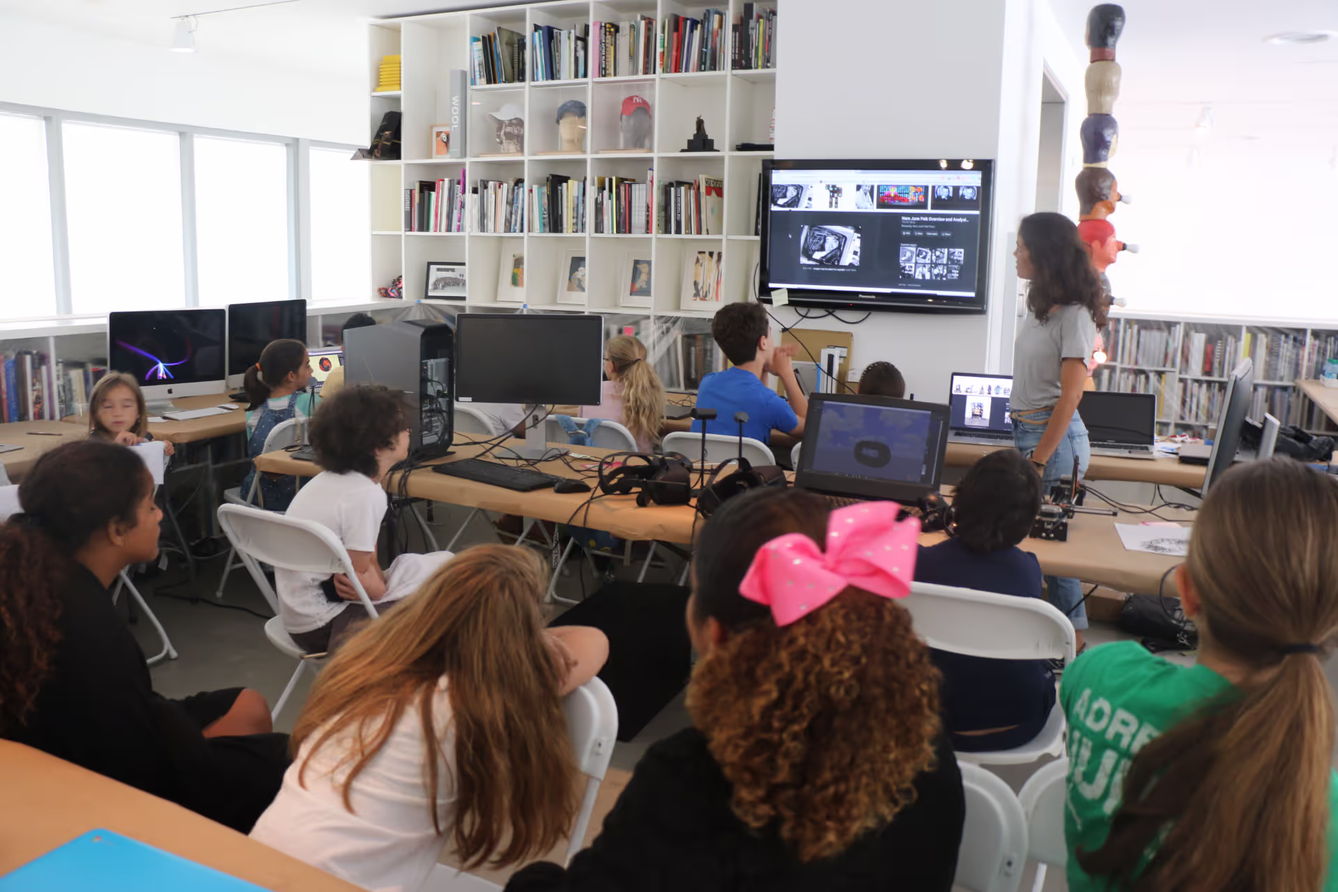 MUD Foundation XRCamp at the De La Cruz Collection in Miami, Florida, 2018, featuring a group of children seated in a classroom while a woman stands near a screen displaying digital images, with computers and VR headsets on tables.