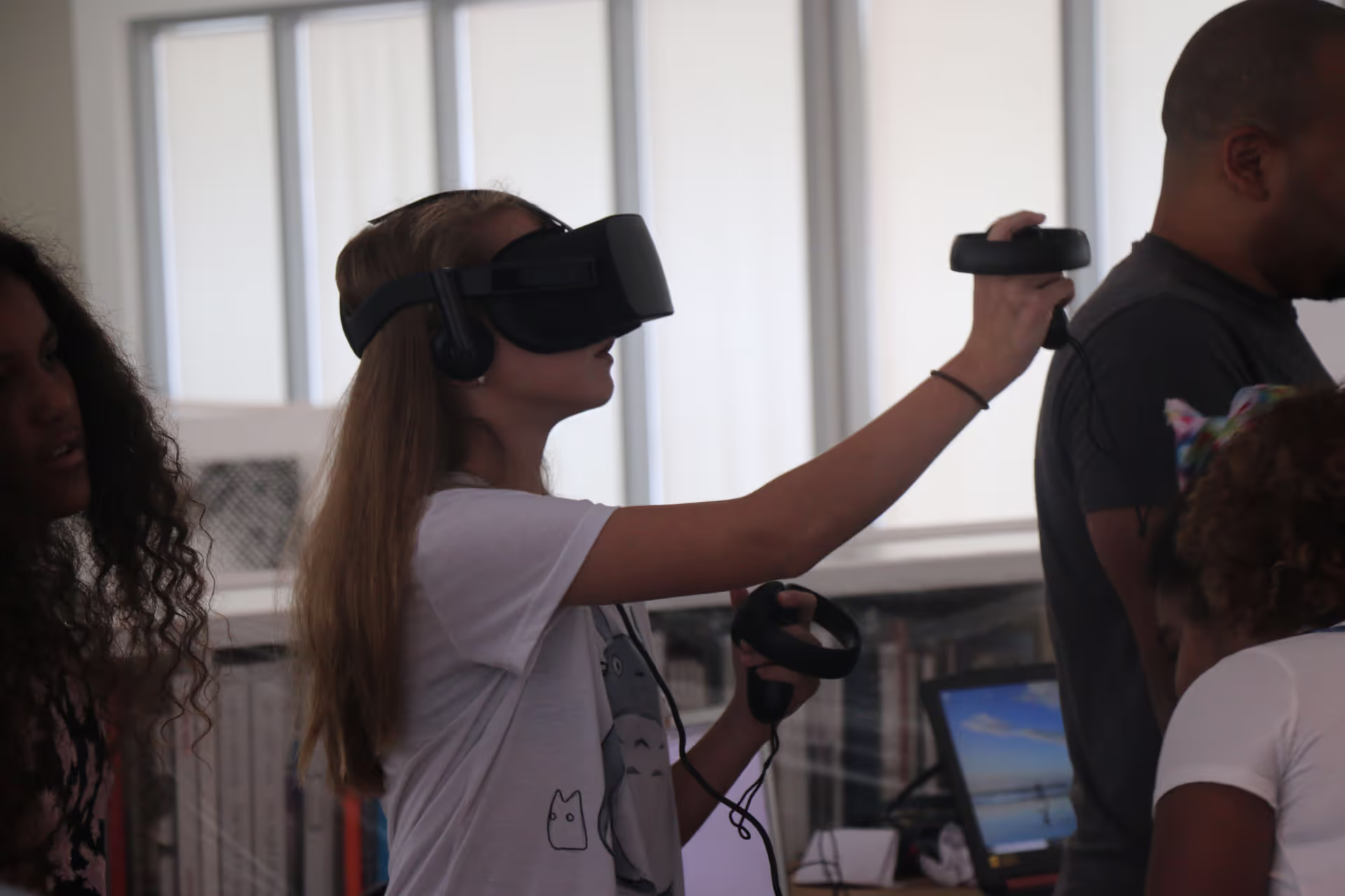 MUD Foundation XRCamp at the De La Cruz Collection in Miami, Florida, 2018, featuring a young woman wearing a VR headset and holding controllers while interacting with a virtual environment, with other participants and a computer monitor visible in the background.