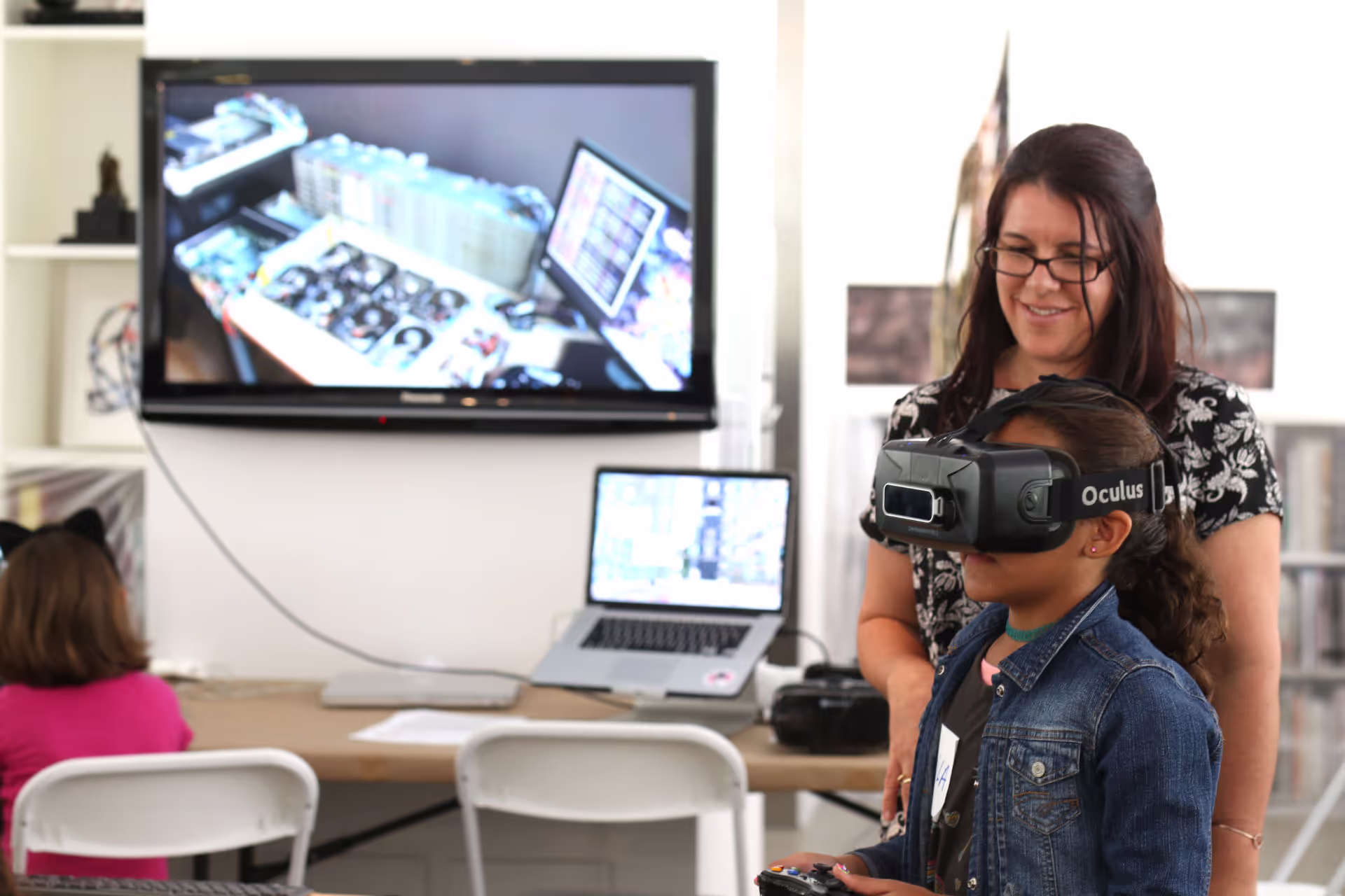 MUD Foundation XRCamp at the De La Cruz Collection in Miami, Florida, 2017, featuring a young girl wearing an Oculus VR headset while a woman watches, with a monitor and laptop displaying related virtual reality content in the background.