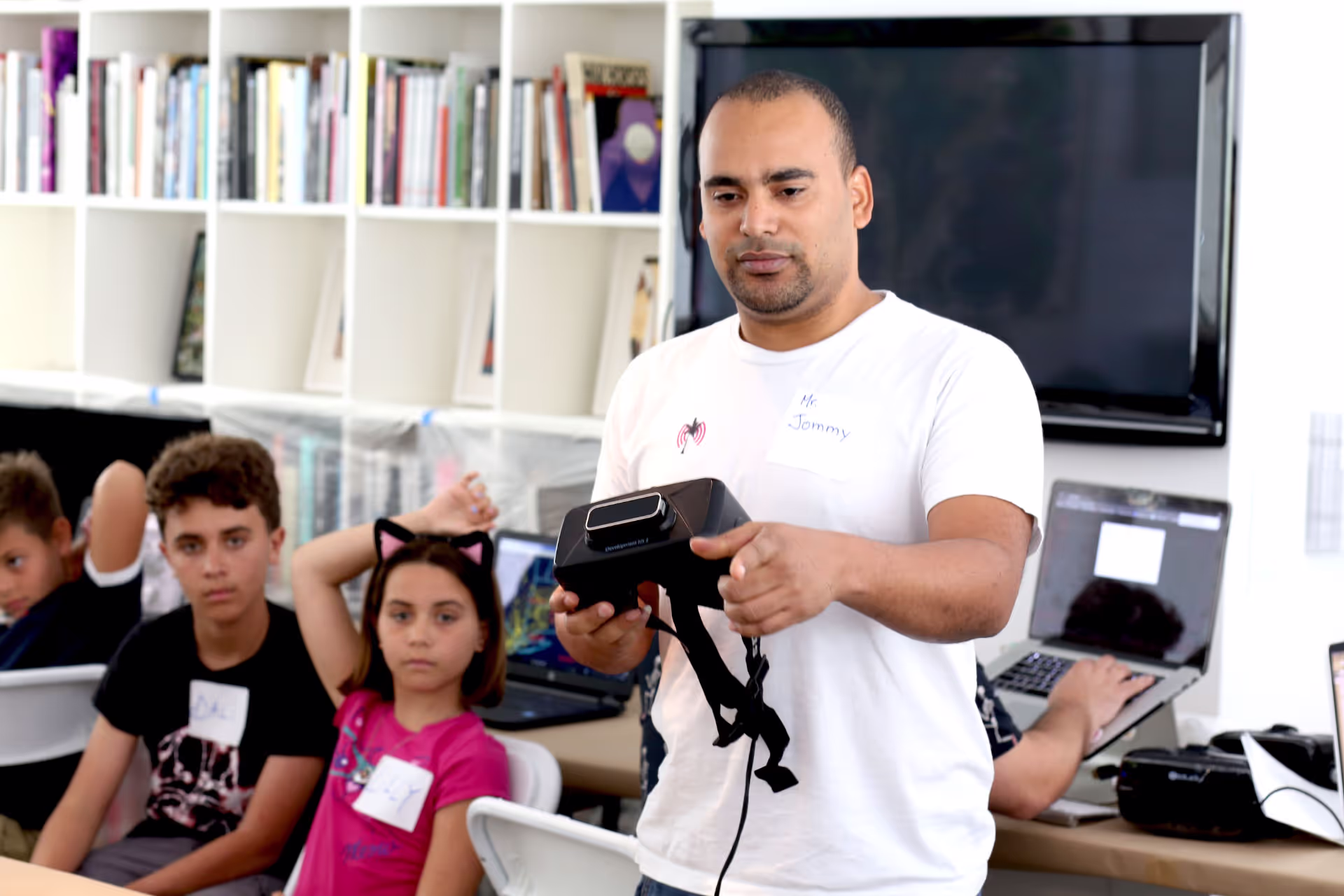 MUD Foundation XRCamp at the De La Cruz Collection in Miami, Florida, 2017, featuring a man holding a virtual reality headset in front of a group of seated children, with bookshelves and laptops in the background.