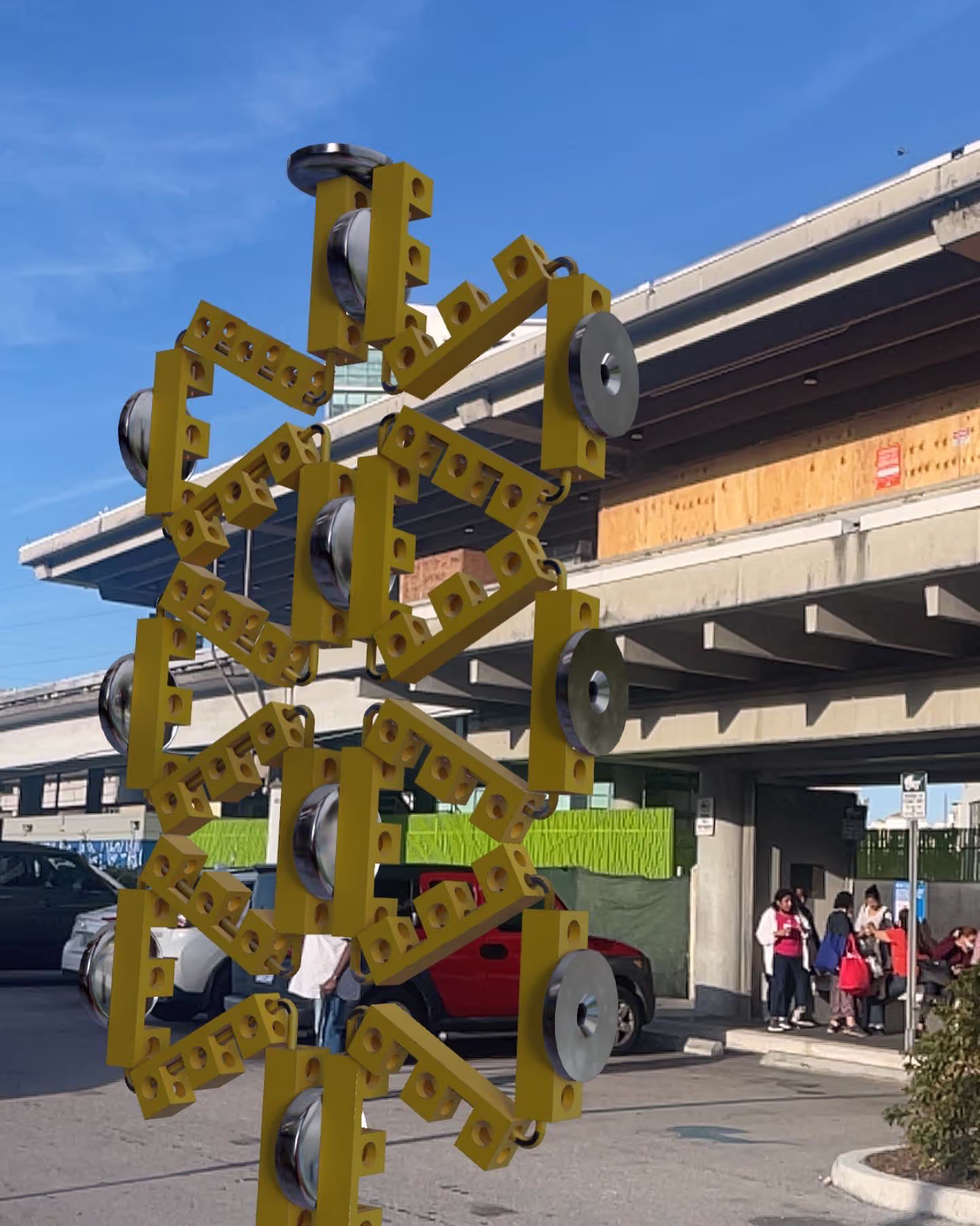 MUD Foundation Rafael Rodríguez Street Activation, featuring an outdoor abstract sculpture made of interconnected yellow metal beams and large metal washers, installed near cars and an overpass with people in the background.