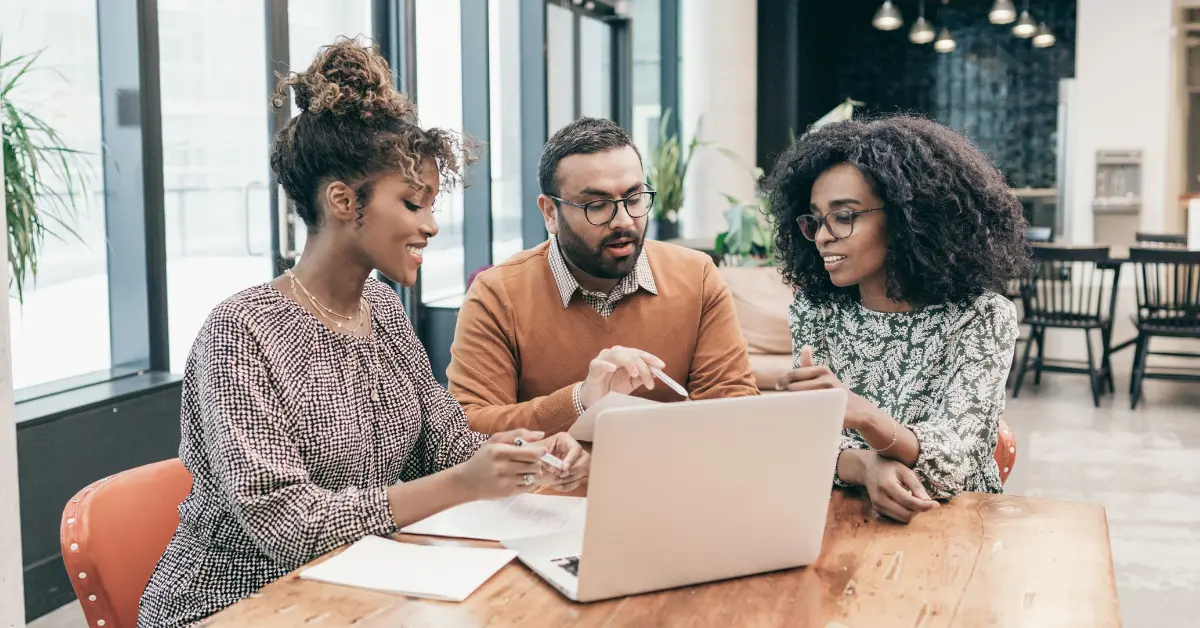 Three professionals collaborating at a table with a laptop, discussing ideas in a modern office setting.
