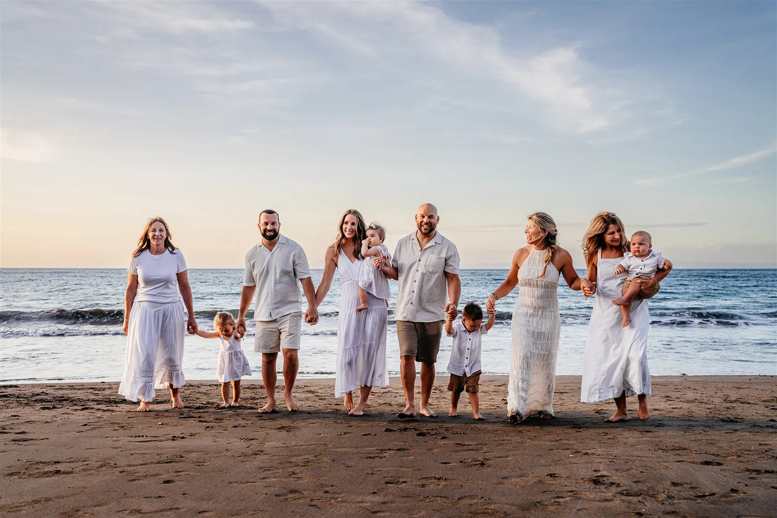 A multigenerational family holding hands and walking barefoot on a beach at sunset.