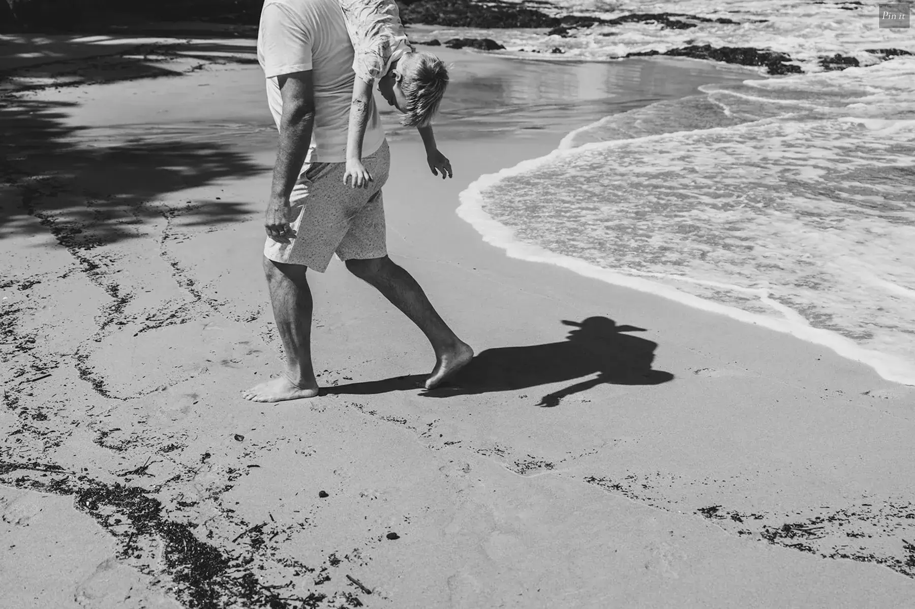 Man carrying a child on his shoulders walking barefoot on a sandy beach near the ocean.