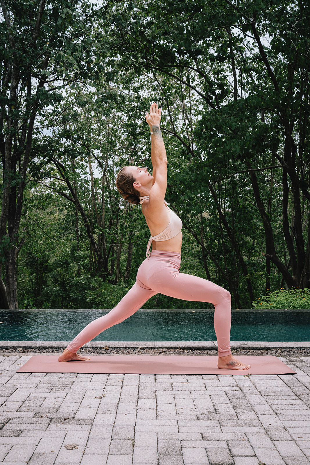 A woman in pink leggings and a beige sports top practices yoga outdoors on a pink mat, performing a high lunge pose with arms raised above her head against a backdrop of green trees and a pool.