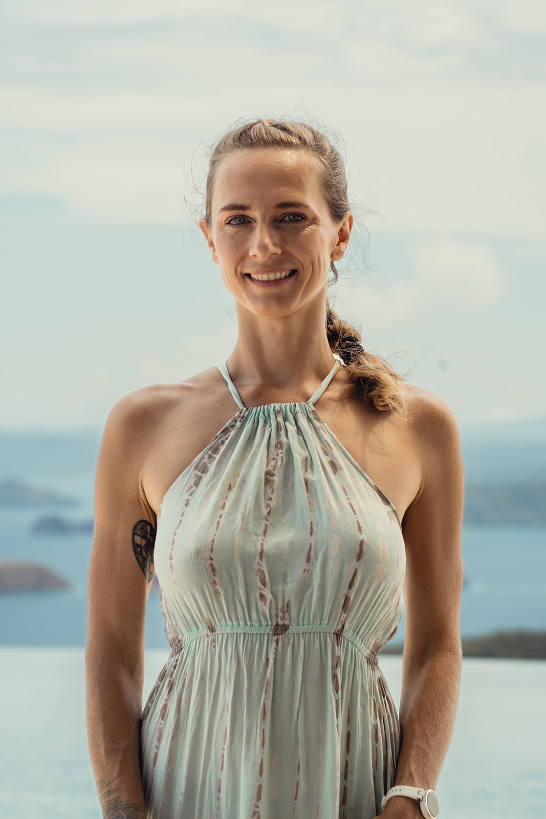 Smiling woman with braided hair wearing shell earrings, a shell necklace, and a white lace top against a soft sky background.