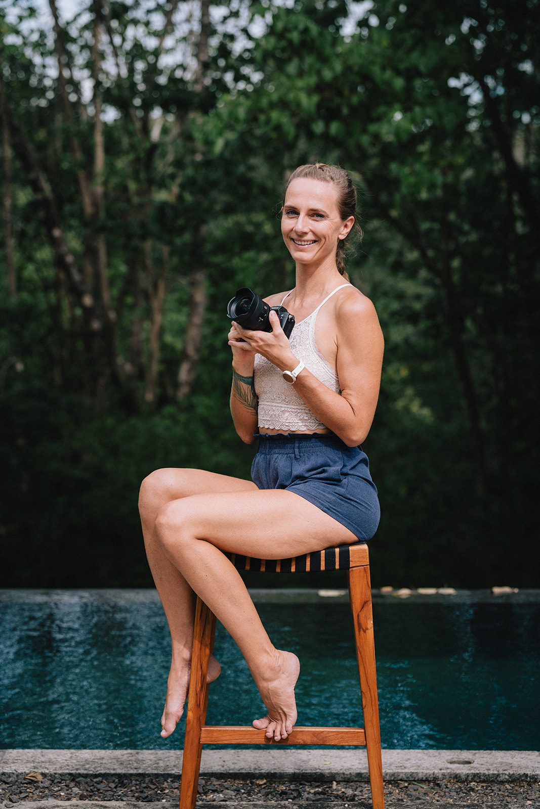 A smiling woman in a lace crop top and blue shorts sitting barefoot on a wooden stool outdoors, holding a camera with a blurred green forest background and a body of water behind her.