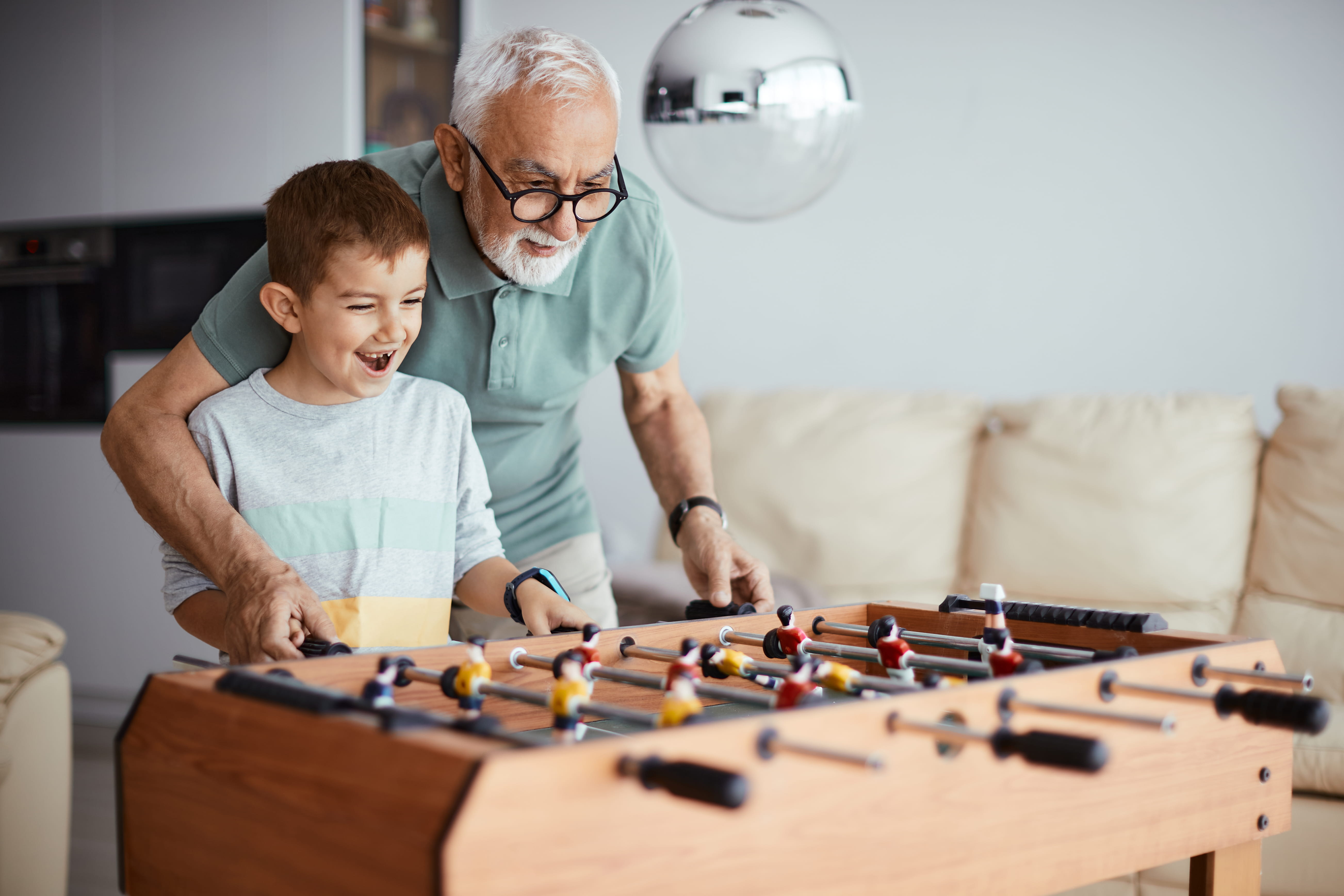 Gray-haired man and young boy happily playing foosball together in a living room.