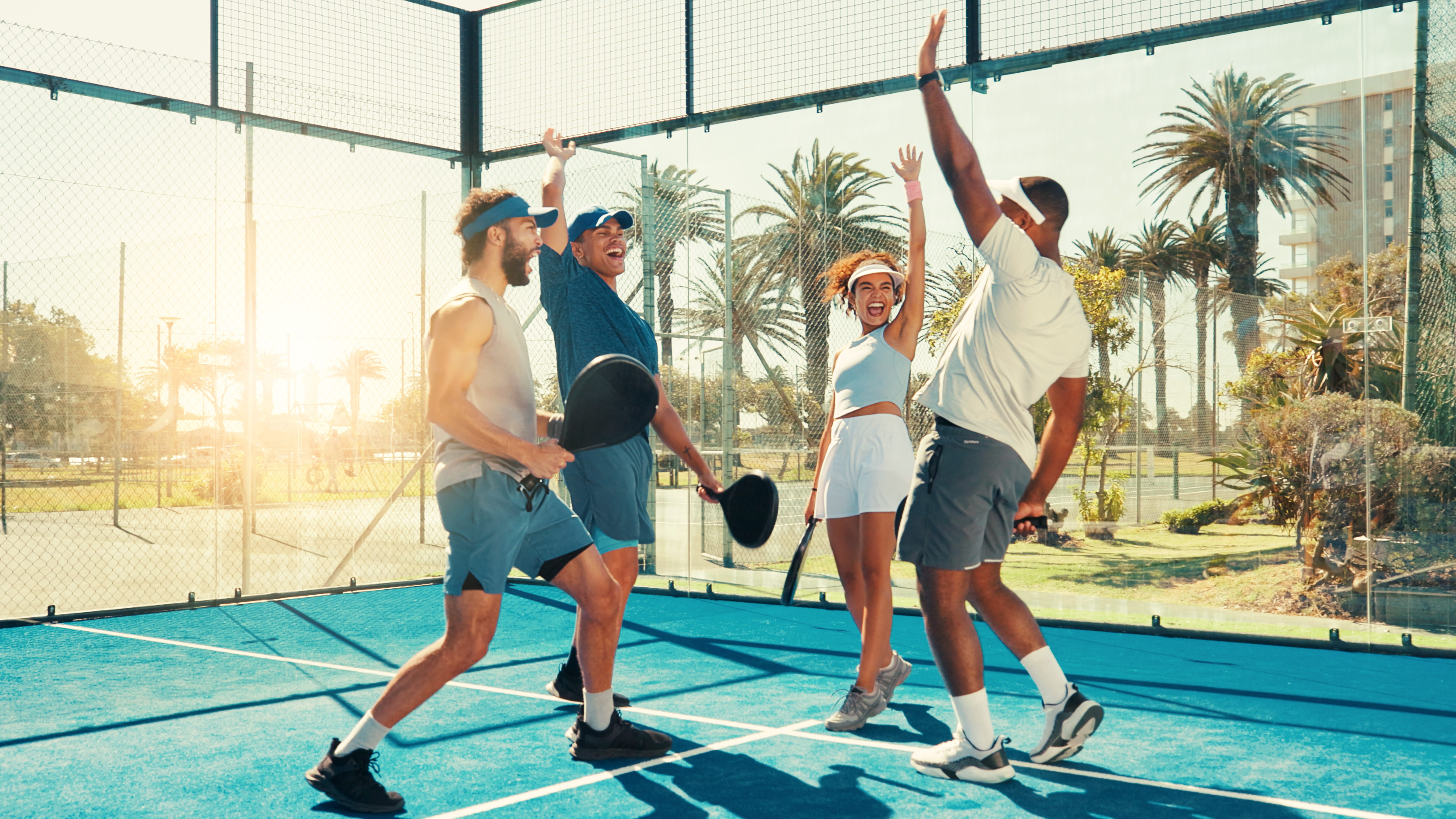 Four friends celebrating winning a paddle tennis game on a bright blue court with palm trees.