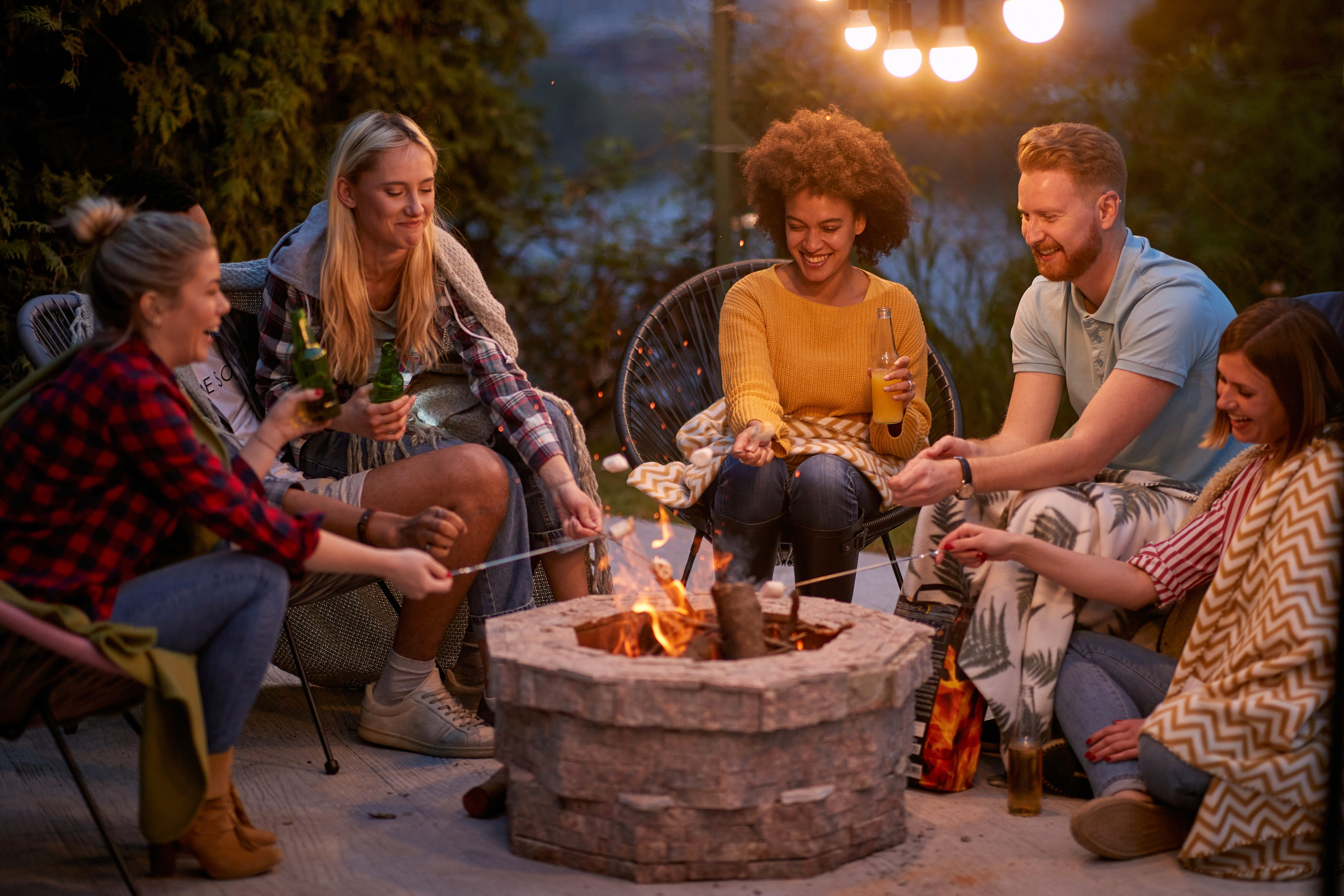 Five adults sitting around a fire pit, roasting marshmallows and holding drinks outdoors at dusk.