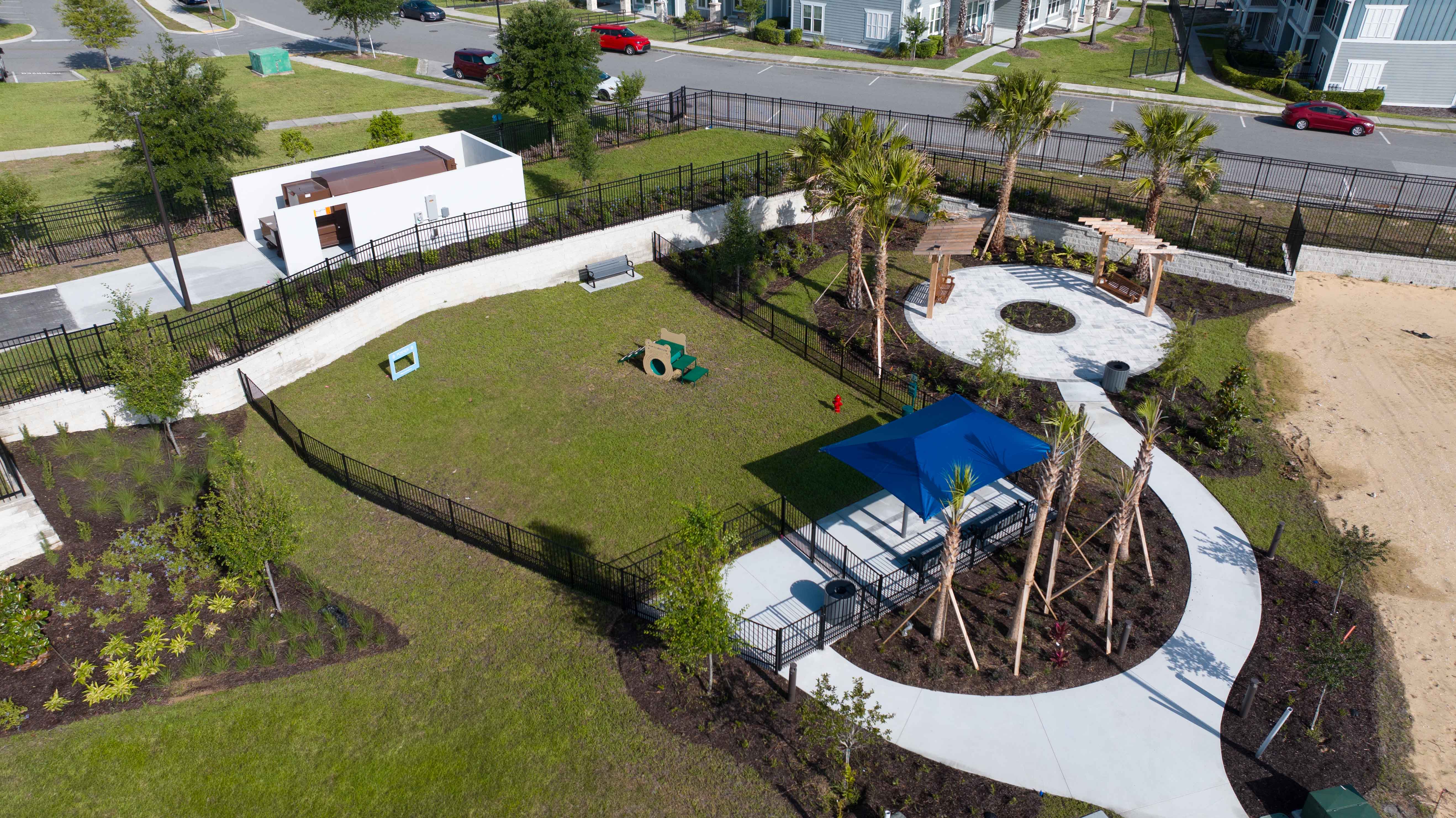 A fenced grassy park area with a small playground, shaded seating, palm trees, and paved walkways.