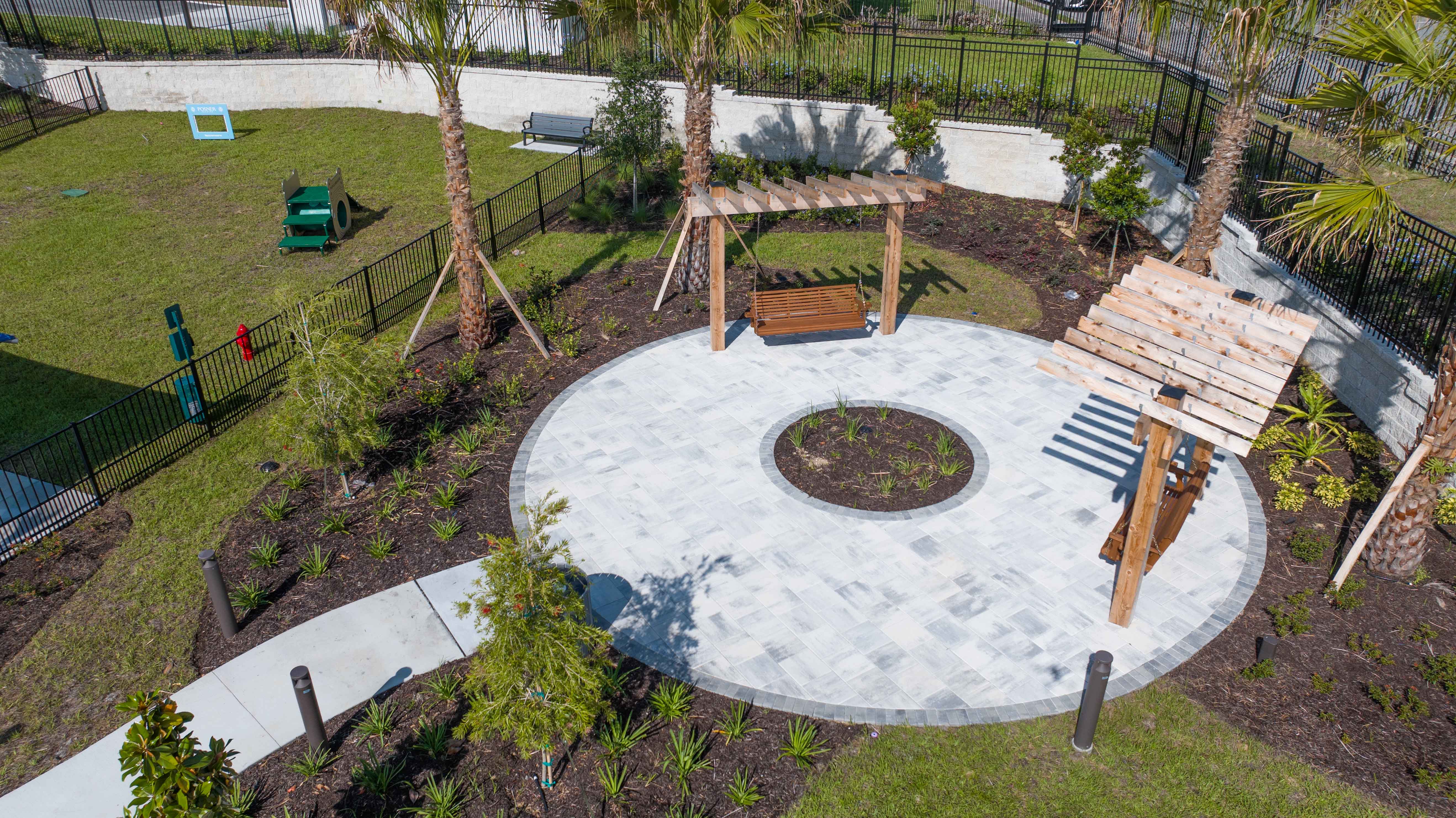 Outdoor garden with circular tiled patio, wooden pergolas, swinging benches, palm trees, and landscaped plants.