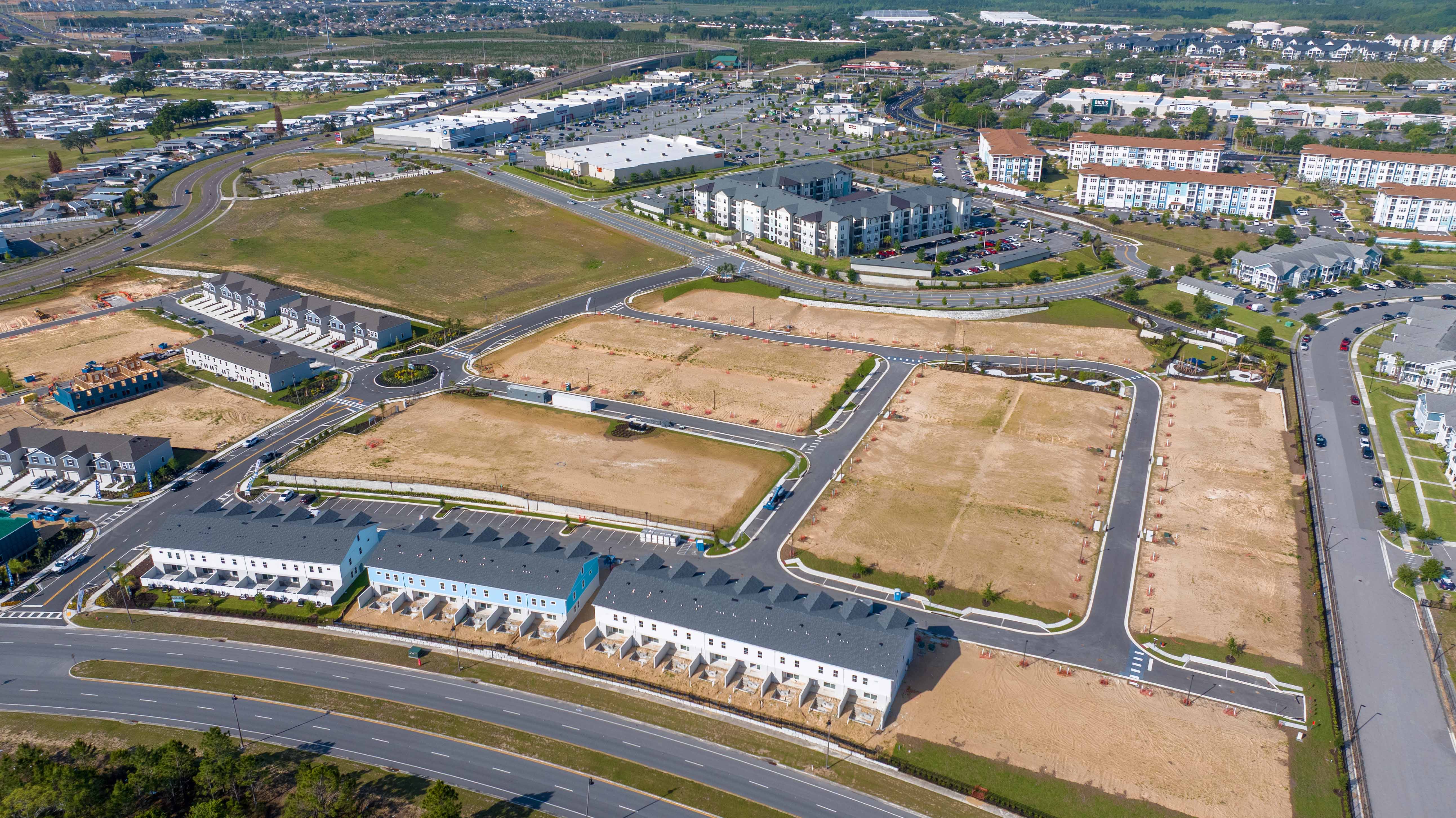 Aerial view of a suburban development with townhouses, empty lots, roads, and nearby commercial buildings.
