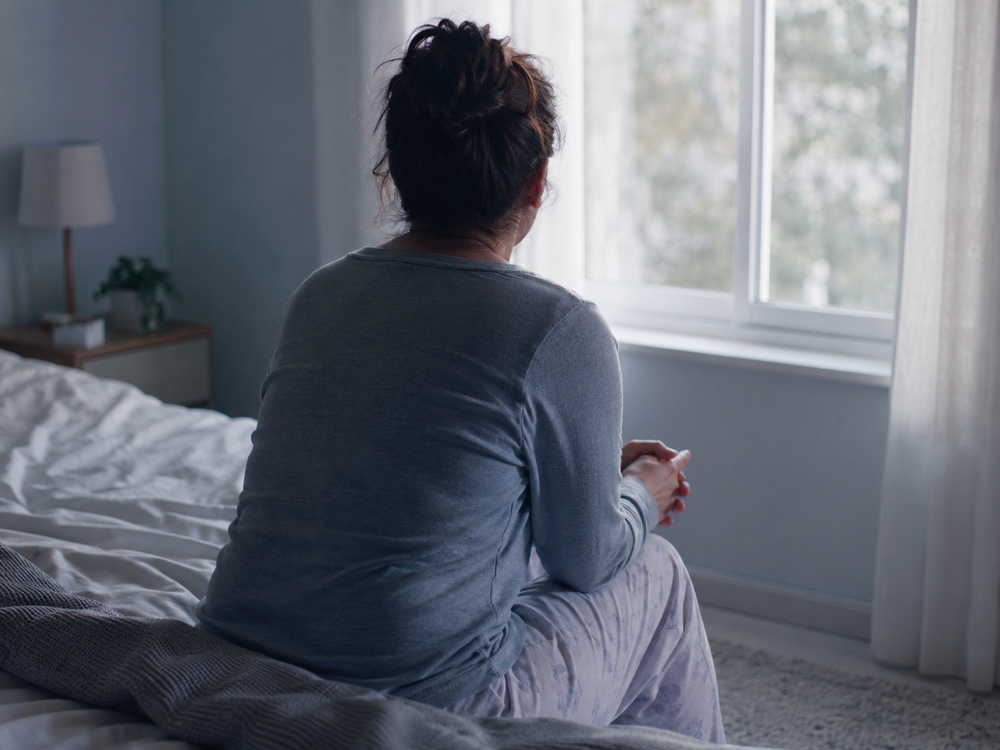 Person sitting on the edge of a bed in soft morning light, hands clasped while looking out the window, conveying discomfort and vulnerability.