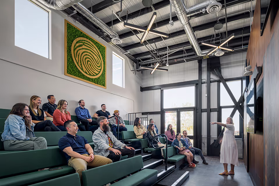 Woman in white dress presenting to an audience seated on green tiered benches in a modern, industrial-style room with large windows and exposed ceiling pipes.
