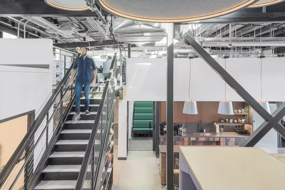 Modern office interior with a blurred man walking down metal stairs, visible kitchen area, and open workspace with exposed ceiling pipes.