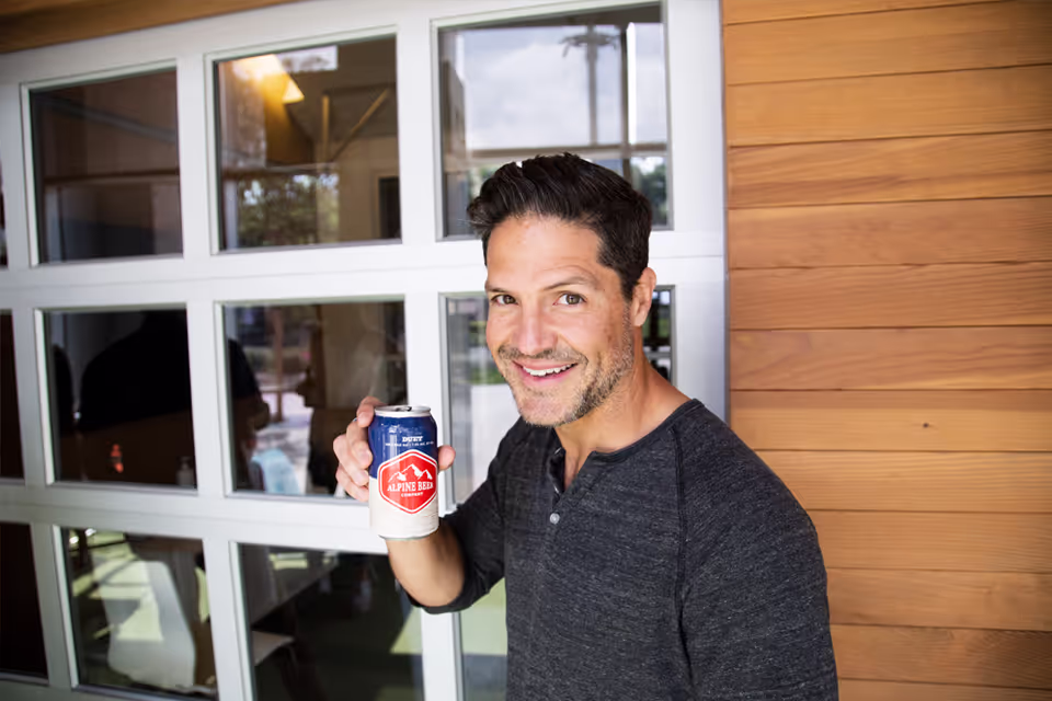 Smiling man holding a can of Alpine Beer with a wooden wall and glass window behind him.