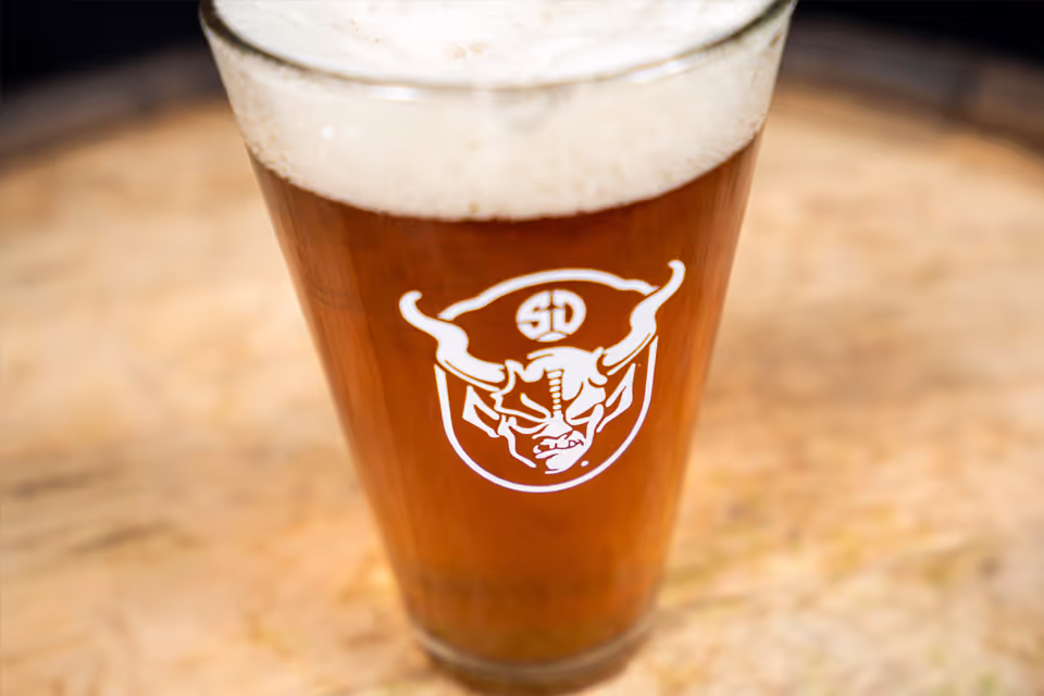 Glass of amber beer with white foam head featuring a logo of a horned skull with 'SD' above it.