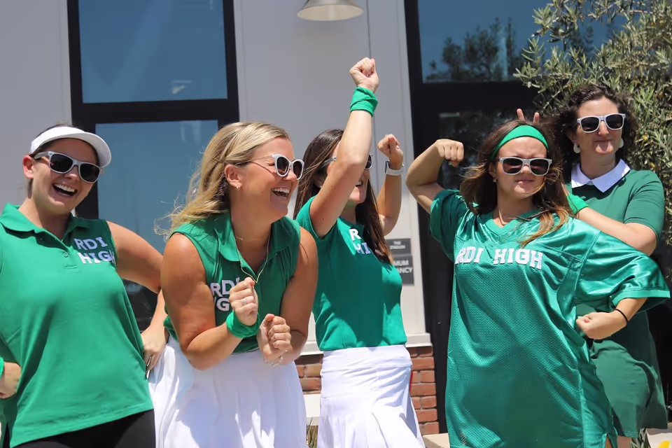 Five women in green RDI High shirts and sunglasses posing outdoors, smiling and flexing their arms in a playful, energetic manner.
