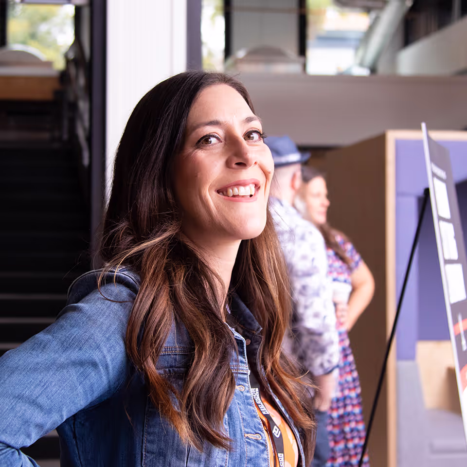 Smiling woman with long brown hair wearing a denim jacket and an event lanyard indoors.