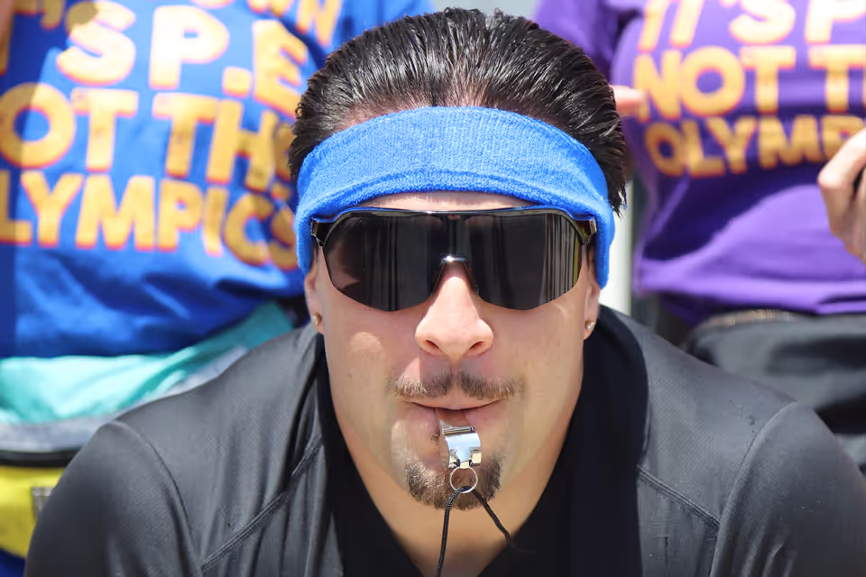 Man wearing a blue headband and black sunglasses blowing a whistle.