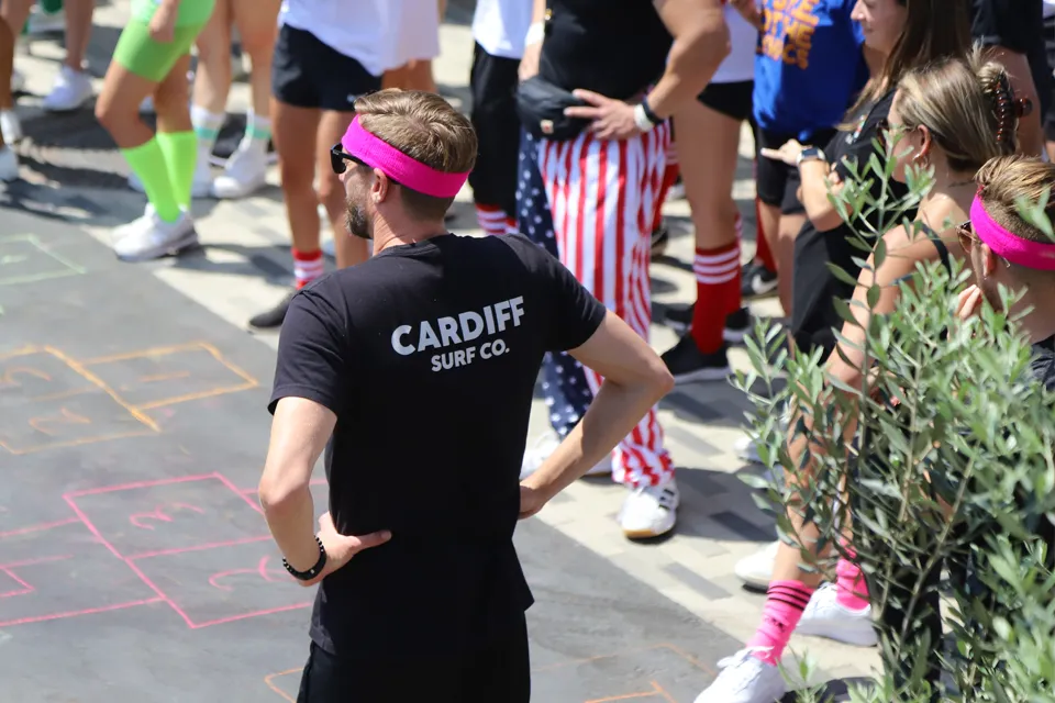 Man wearing a black Cardiff Surf Co. t-shirt and pink headband standing with hands on hips among a crowd dressed in colorful athletic wear and striped pants.