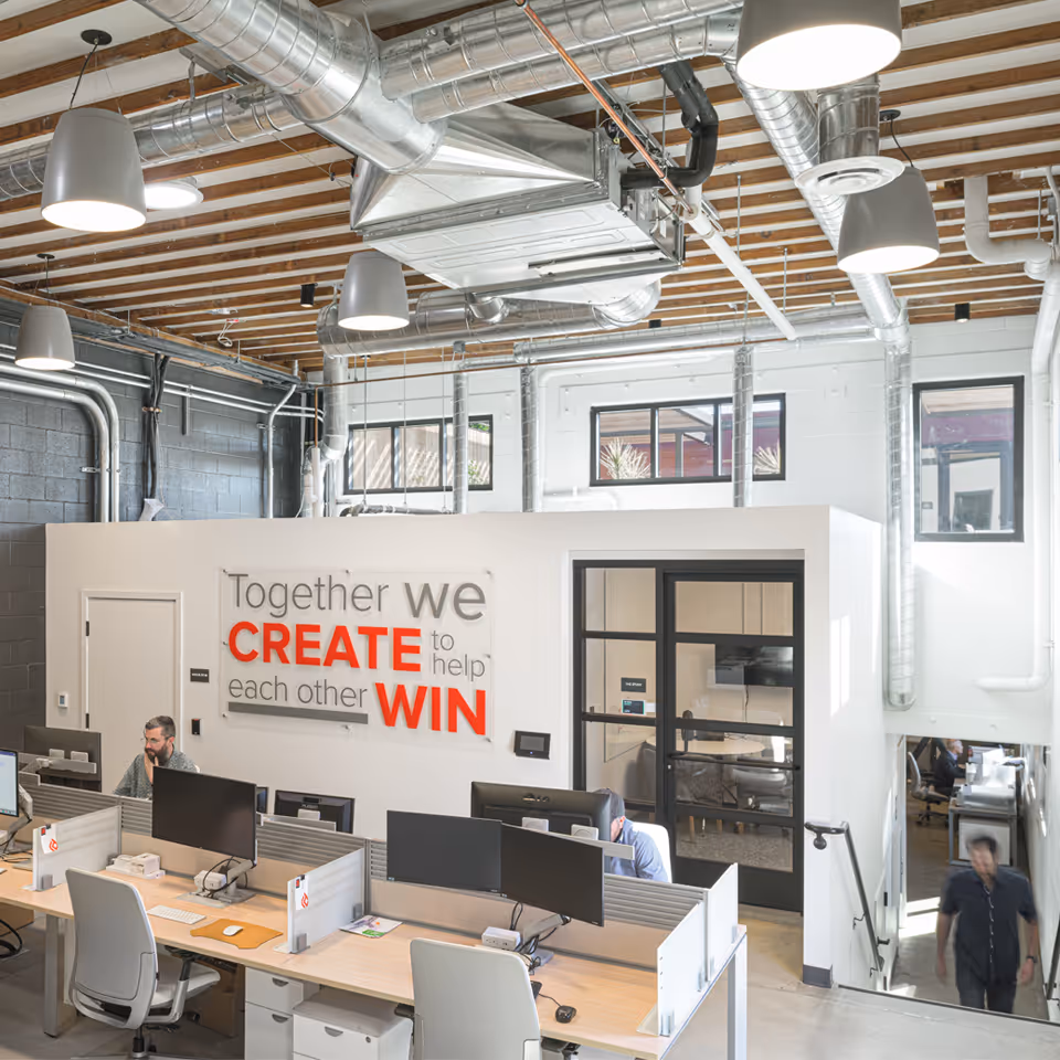 Modern office space with workstations, overhead industrial lighting and ductwork, and a motivational wall sign reading 'Together we CREATE to help each other WIN'.