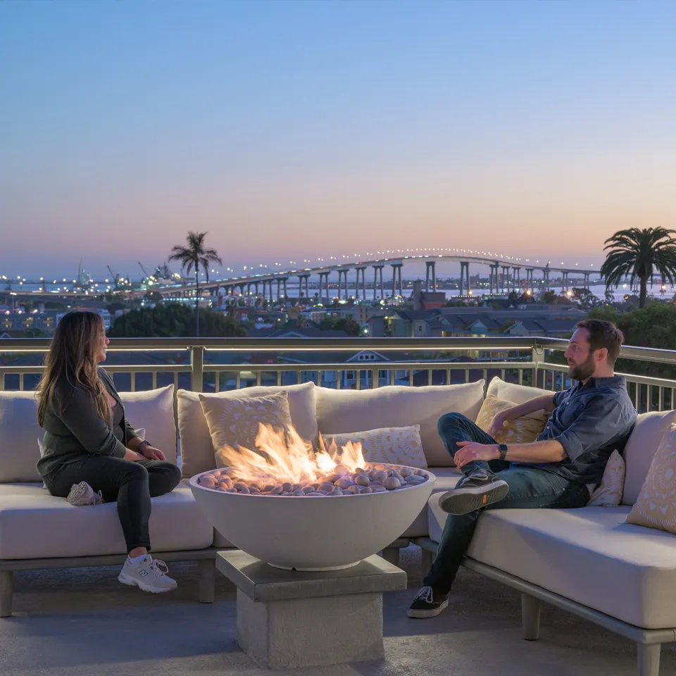 Two people sitting on an outdoor sofa around a lit fire pit with a city bridge and palm trees in the background at dusk.