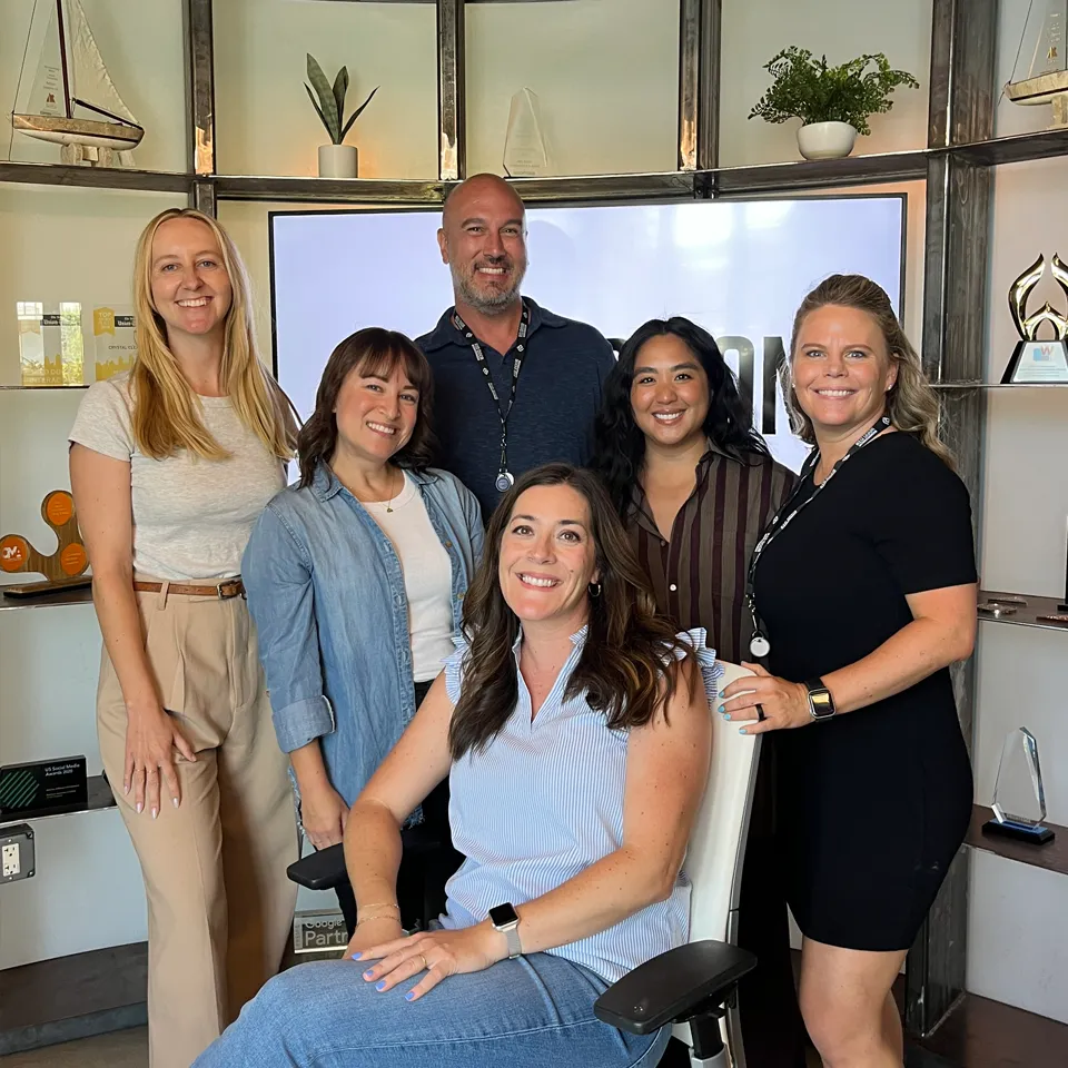 Group portrait of five women and one man smiling in an office setting with awards and plants on shelves behind them.